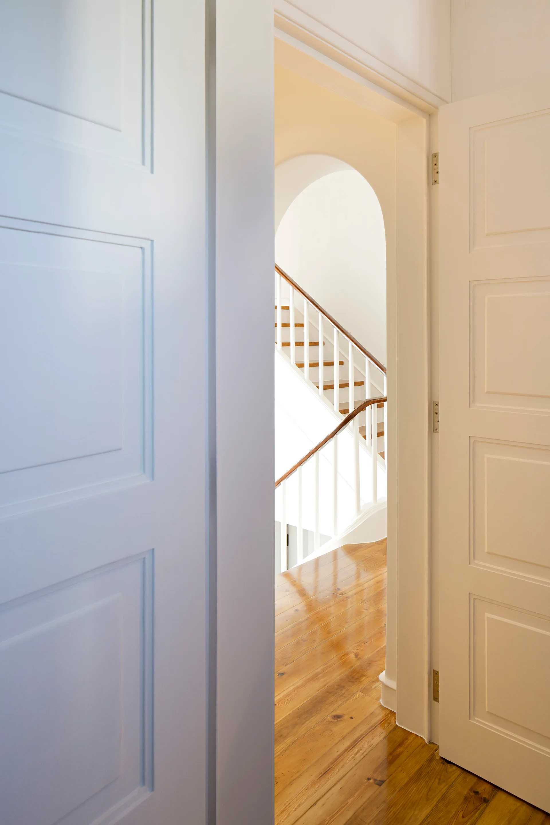 A view through open white paneled doors reveals a polished hardwood hallway floor and a classic staircase framed by an interior archway.