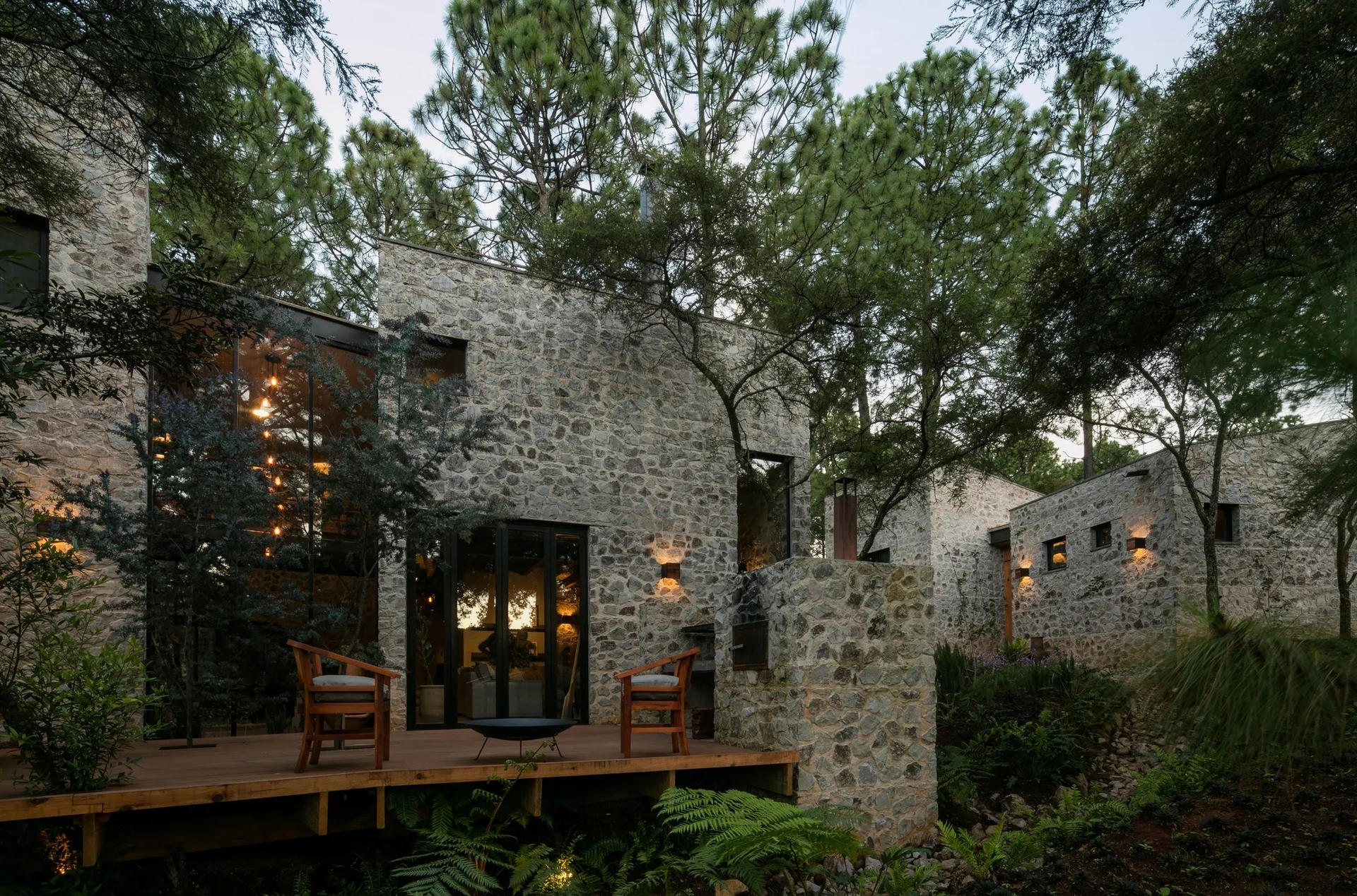 An elevated wooden deck with two chairs and a fire pit sits outside a modern stone cabin surrounded by a dense pine forest.