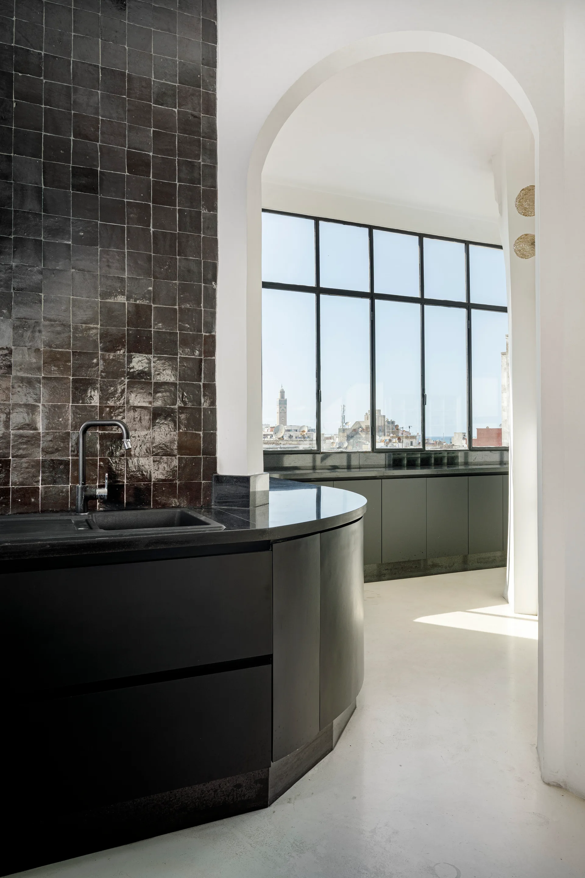 A modern kitchen featuring curved matte black cabinetry and a glossy black tiled backsplash is visible through a clean white plaster archway.