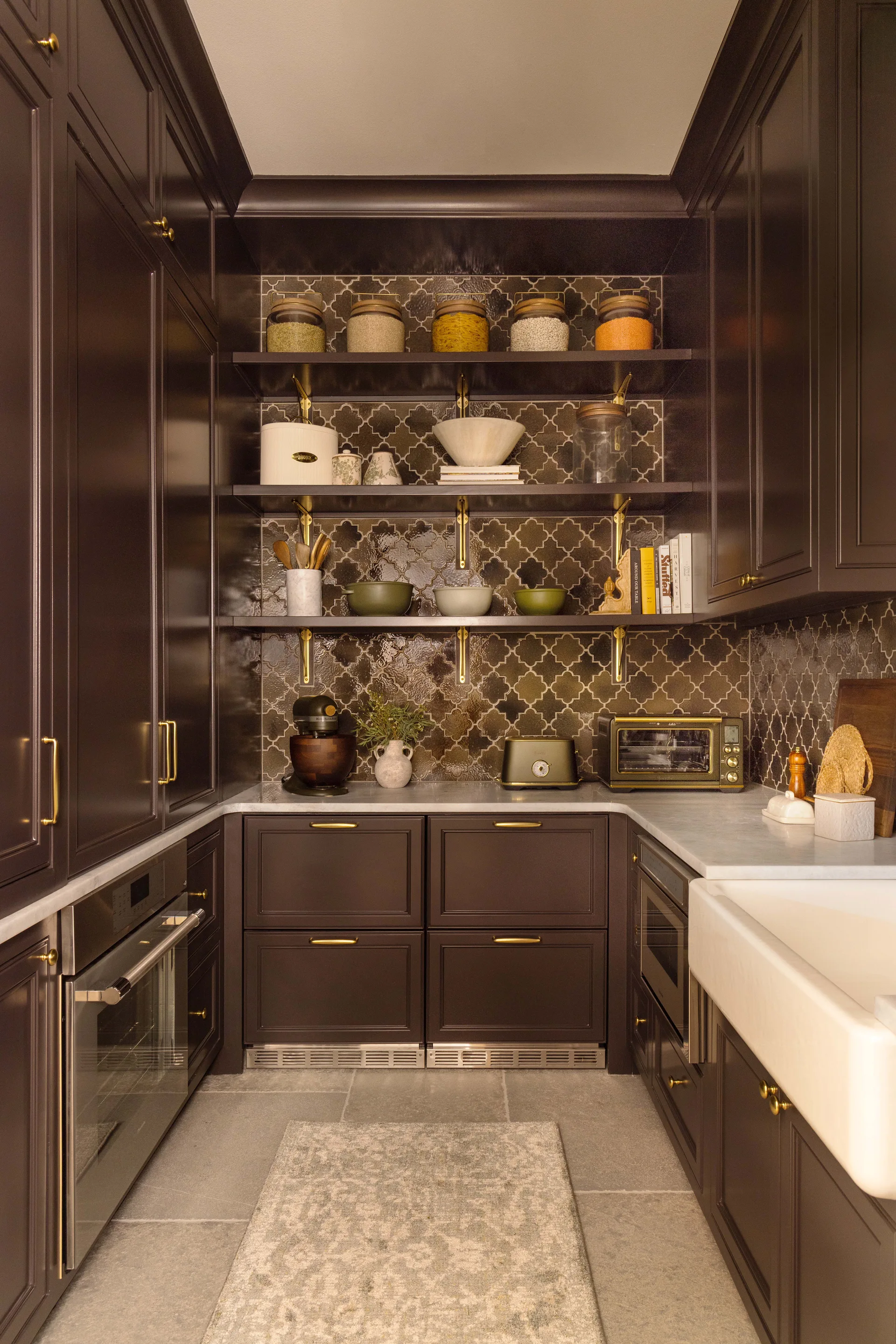 A dark brown u-shaped scullery featuring a white farmhouse sink, brass hardware, and open wooden shelving against a patterned quatrefoil tile backsplash.