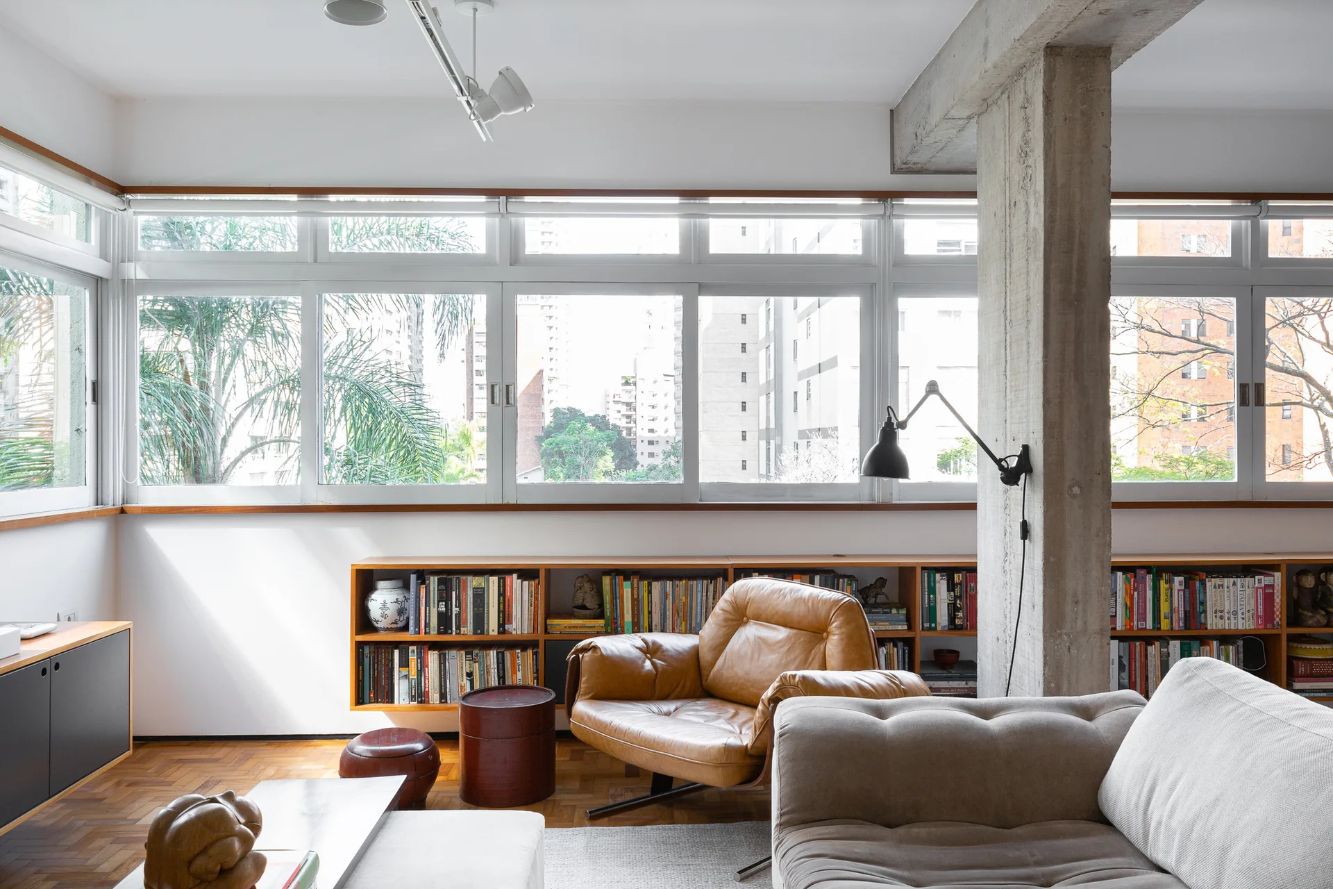 A bright living room with a tan leather armchair, low wood bookshelves under a band of windows, and an exposed concrete pillar.