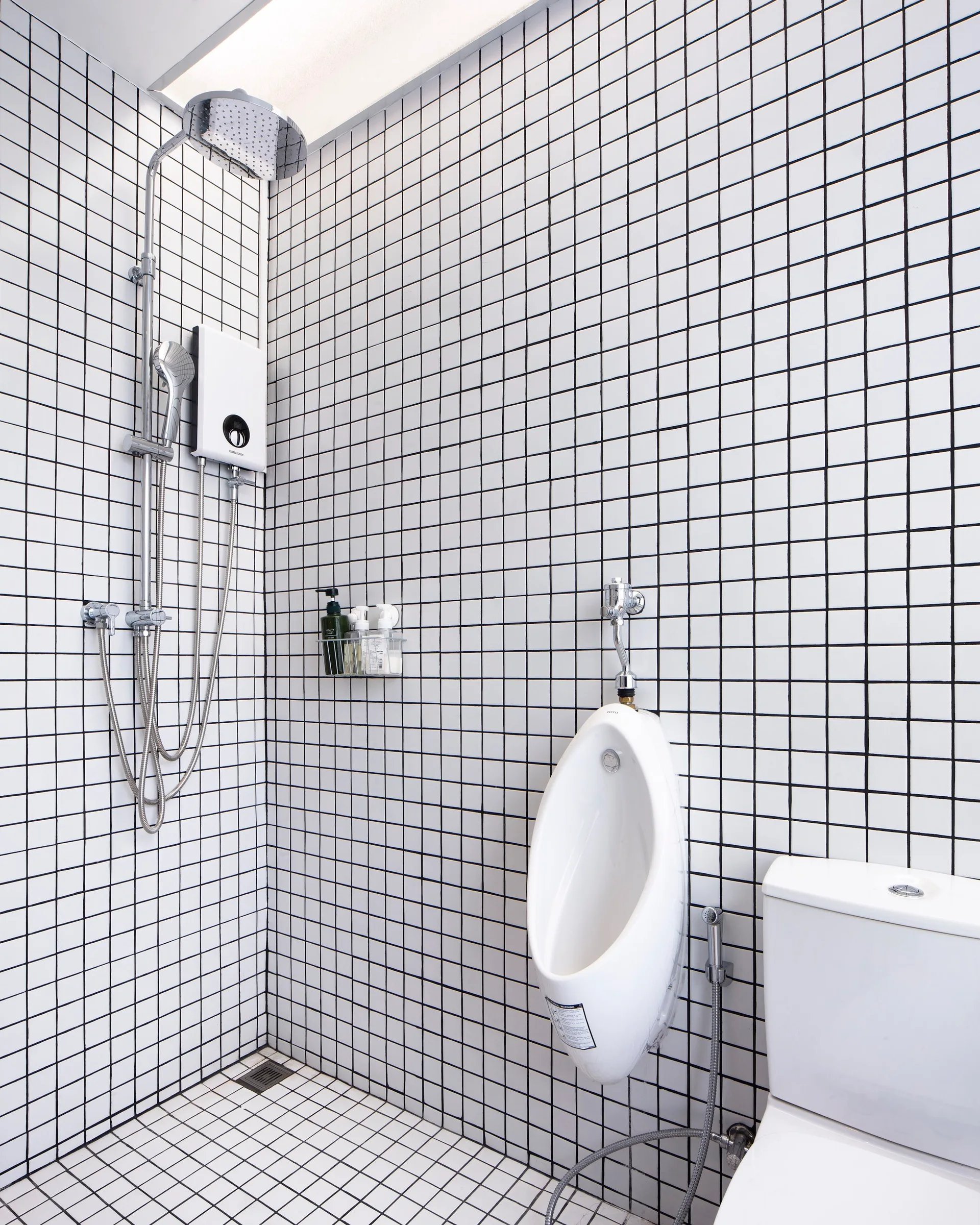 A compact bathroom featuring white square tiles with high-contrast black grout on the walls and floor, complete with a shower, urinal, and toilet.