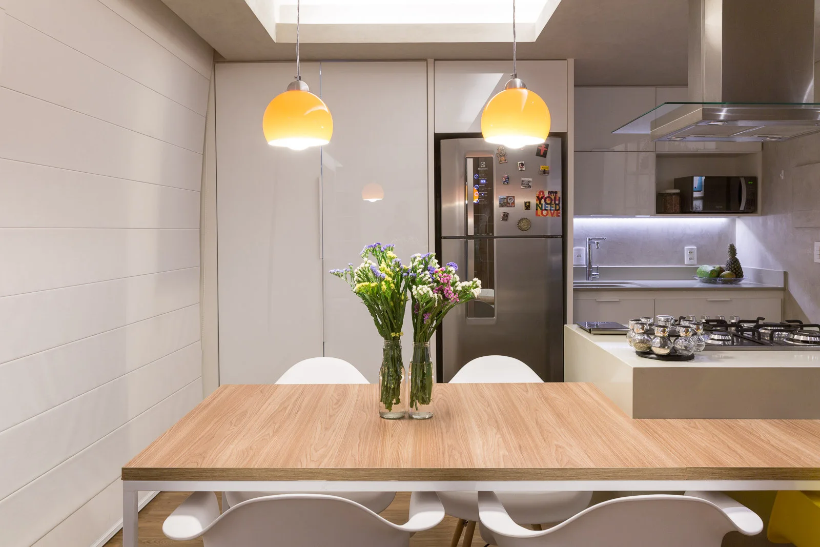 A modern kitchen and dining space featuring a wood-topped table and white chairs under two amber glass pendant lights.