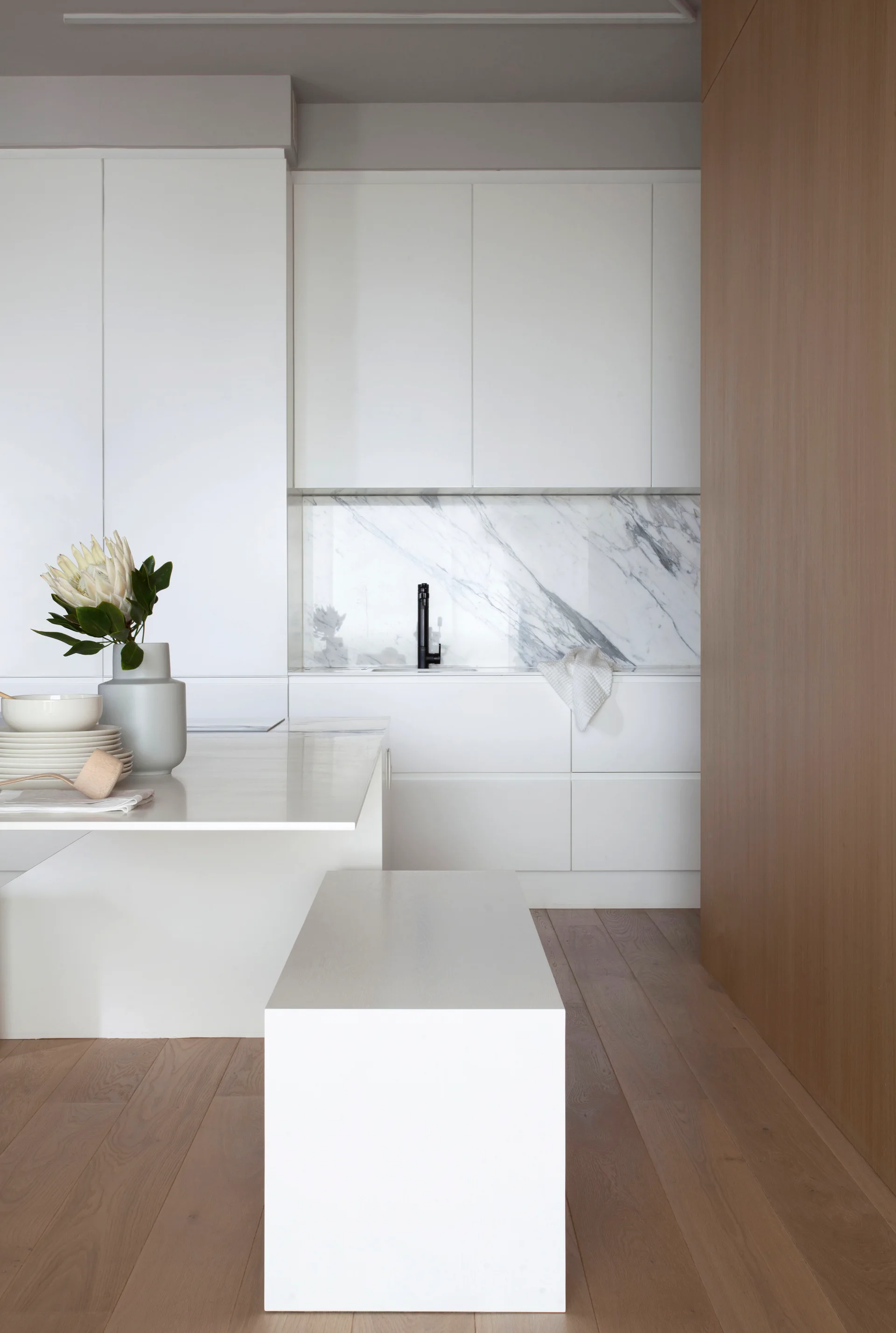 A minimalist kitchen with matte white cabinetry, a veined marble backsplash, a black faucet, and a white bench over light wood floors.