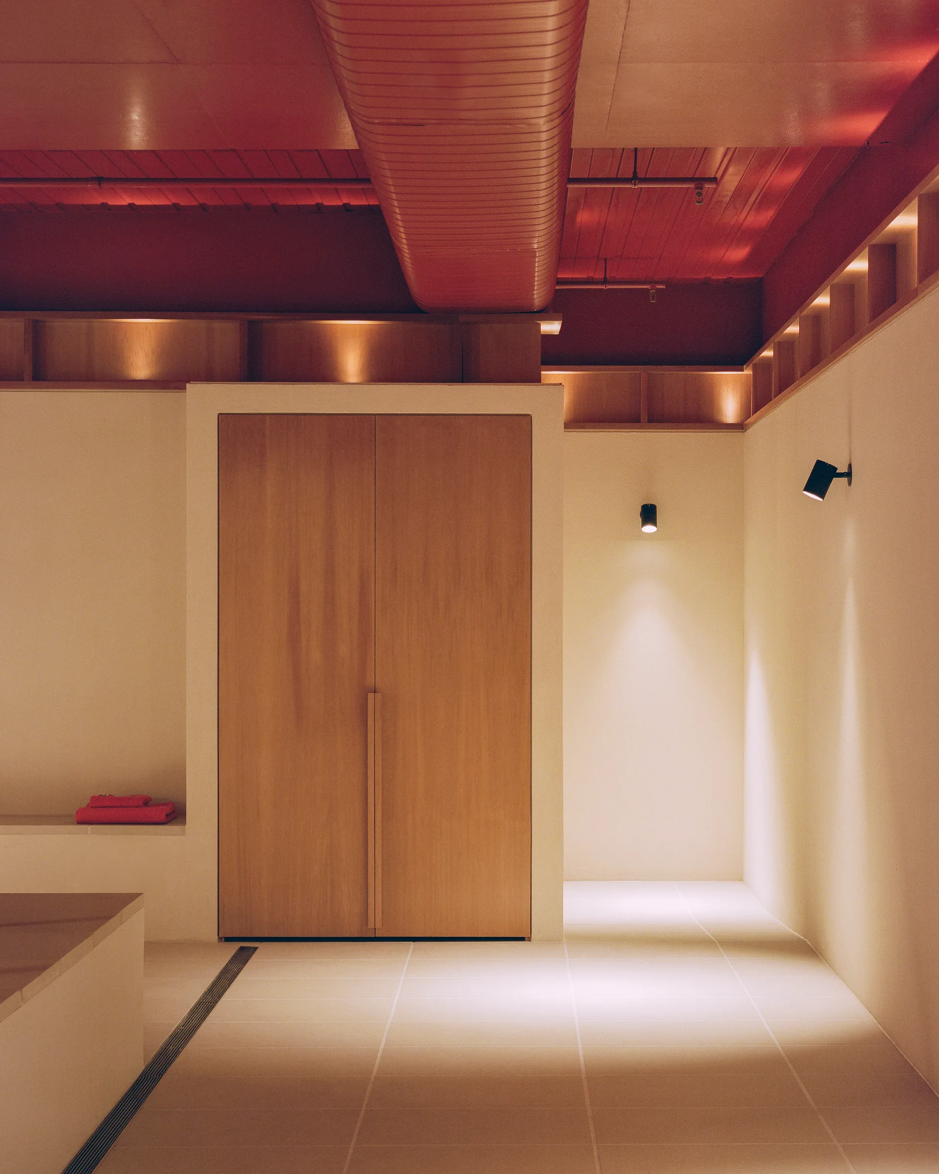 A minimalist spa space featuring off-white tiled floors, a tall light wood cabinet, and a bold red painted ceiling with industrial ductwork.