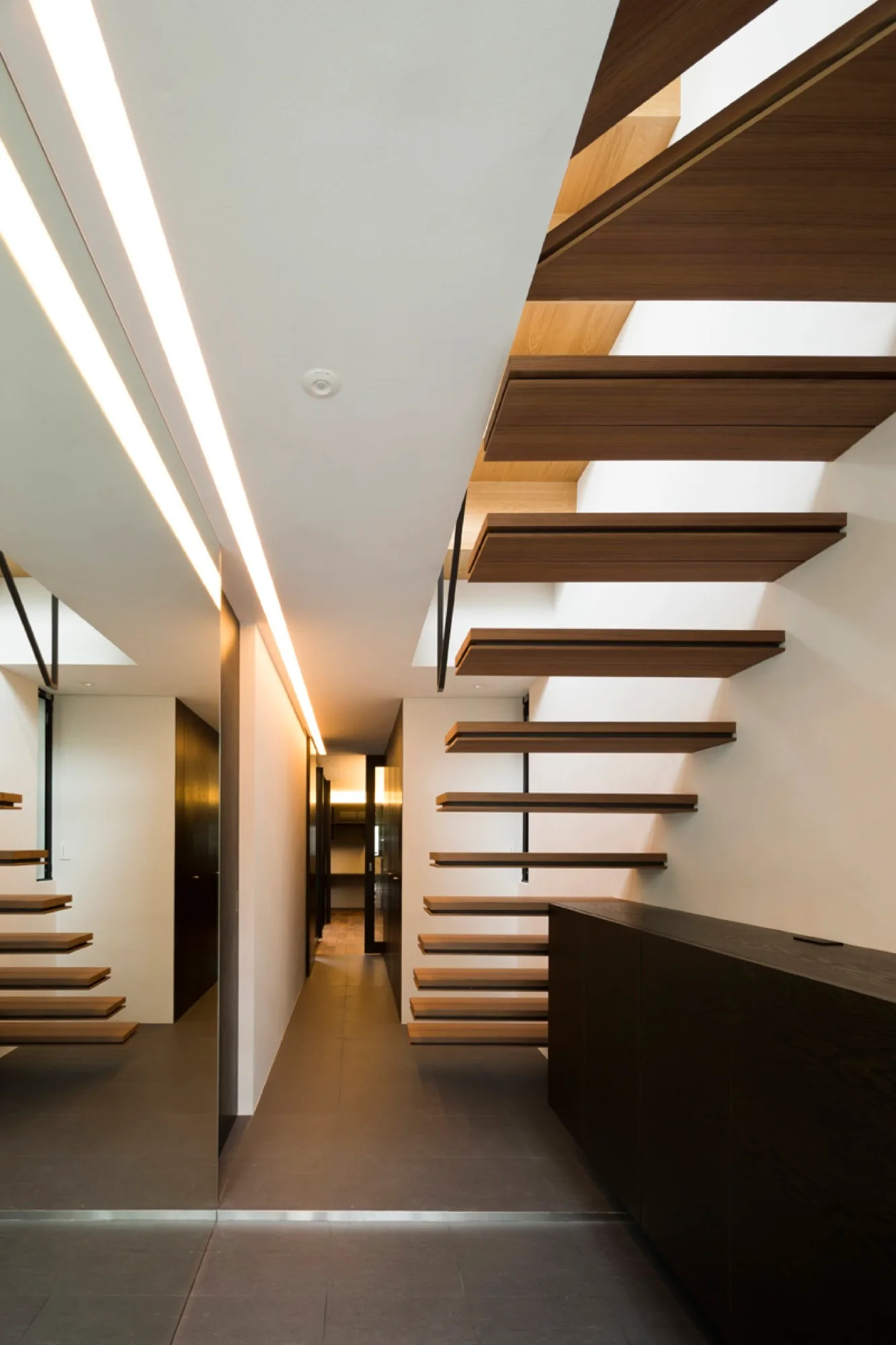 A minimalist hallway with floating wood stairs, a dark low console cabinet, a large wall mirror, gray floor tiles, and ceiling strip lighting.