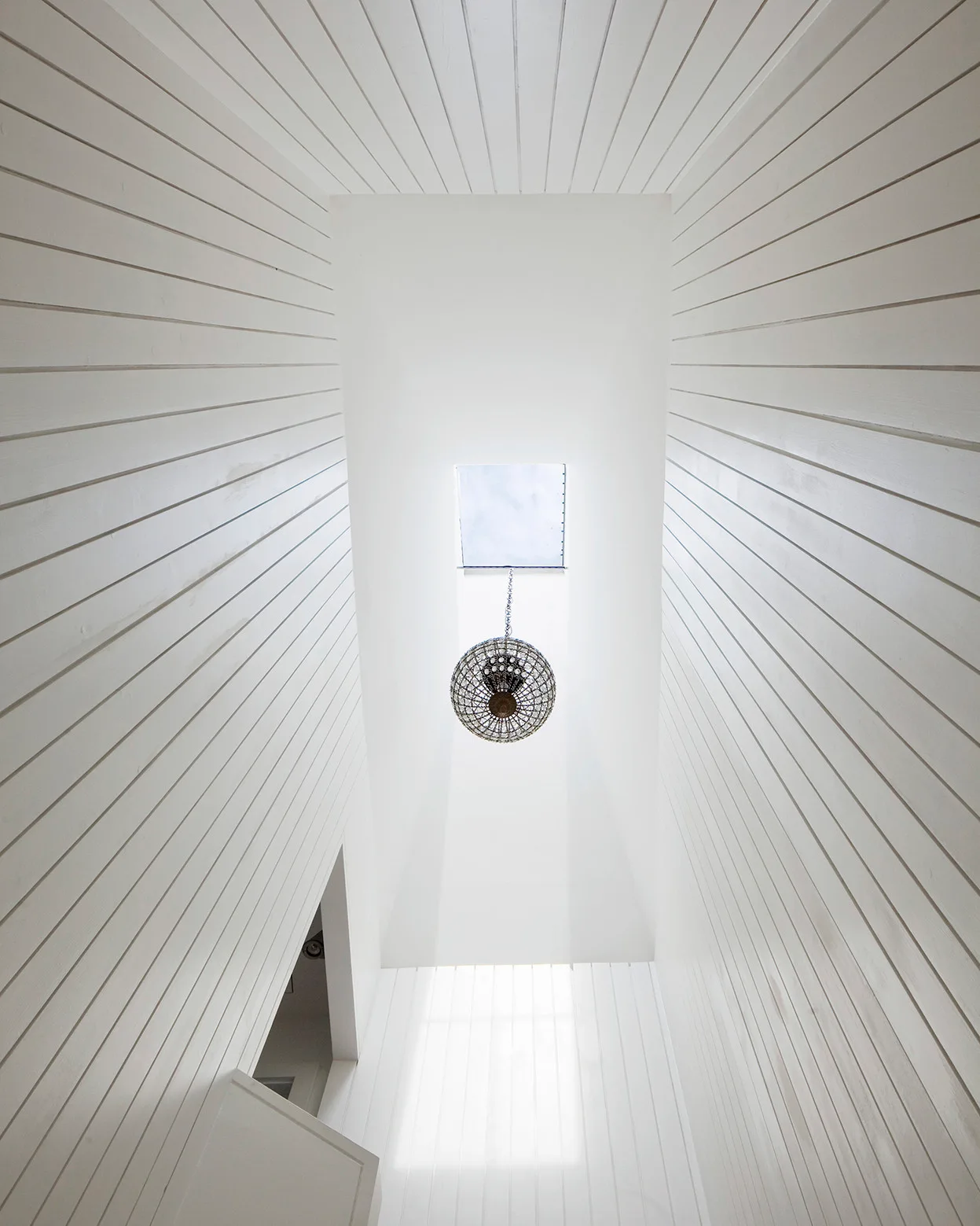 An upward vertical view of a high-ceilinged white shiplap stairwell featuring a central sphere pendant light below a square skylight.
