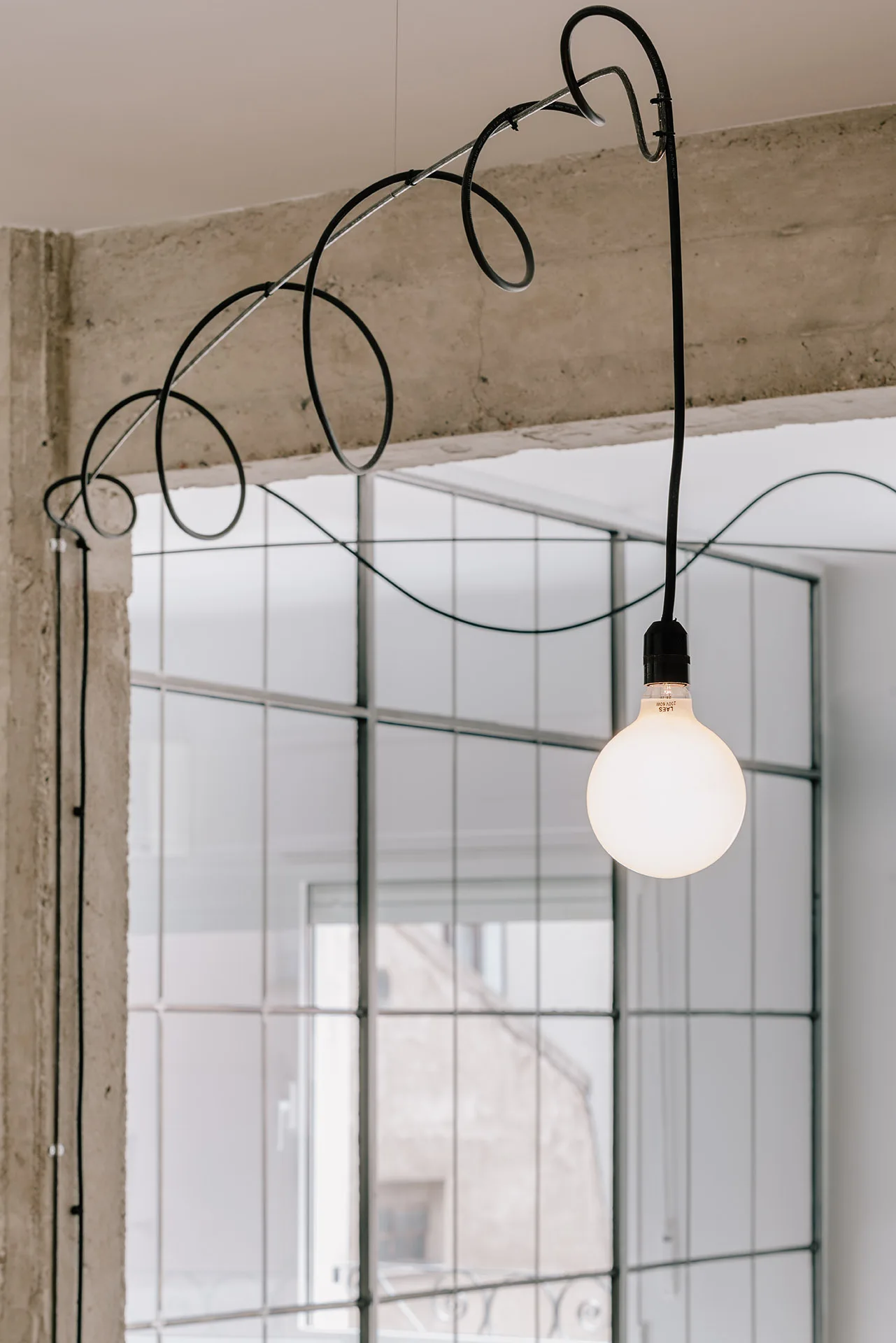 A single glowing globe pendant light hangs from a black cord coiled around a concrete beam in front of a metal grid window partition.
