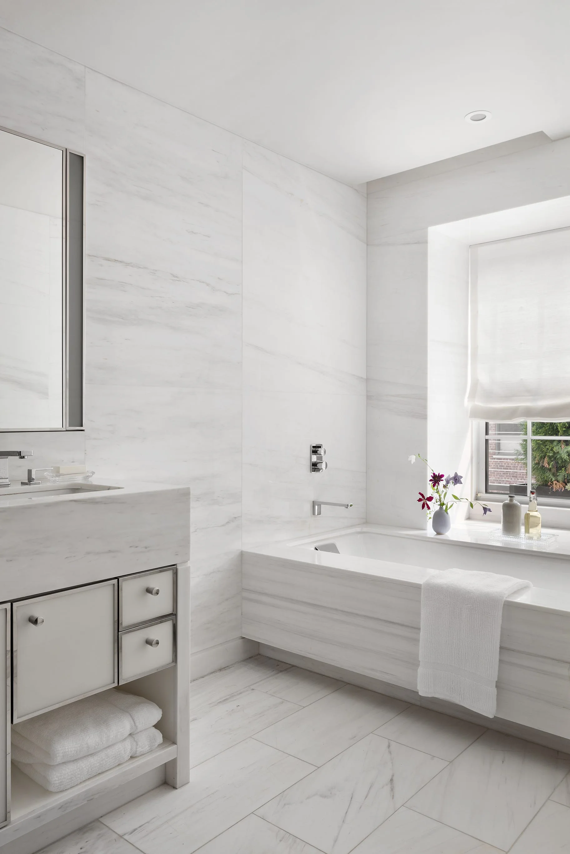 A minimalist bathroom features veined white marble walls and flooring, a built-in bathtub under a window, and a matching stone vanity.