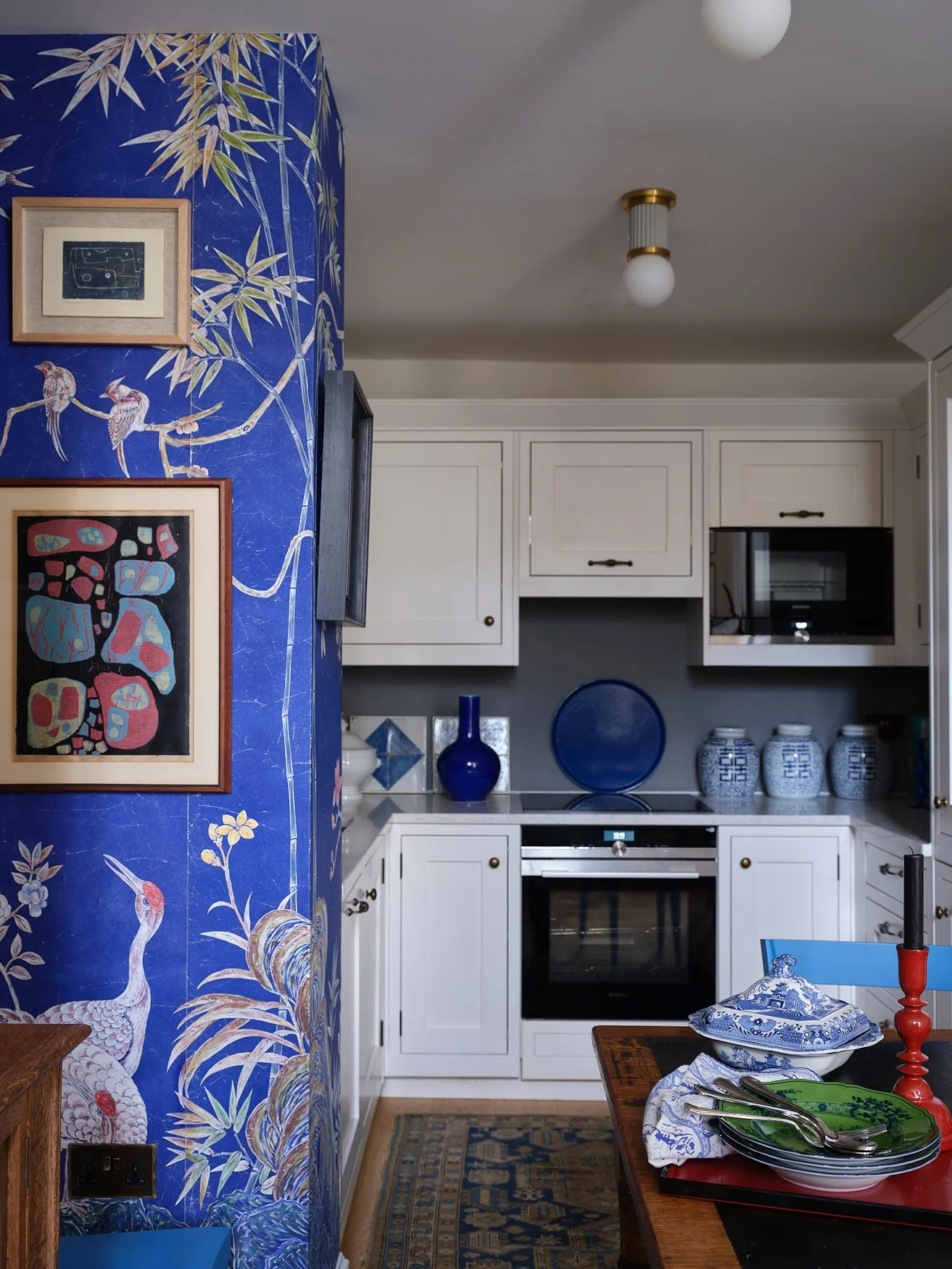 A white kitchen features a structural wall covered in blue bird-patterned wallpaper next to a wood dining table.