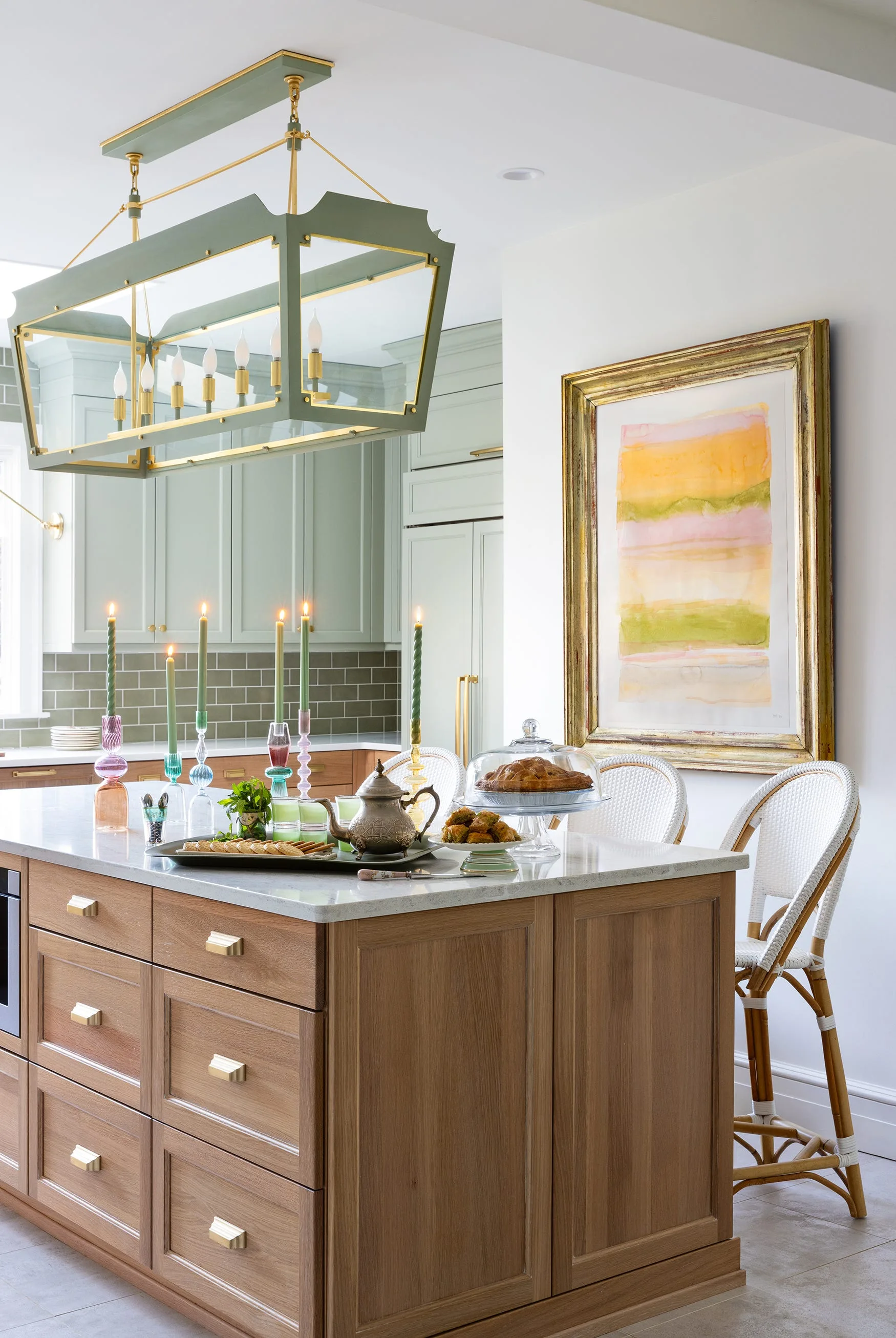 A wooden kitchen island with white woven barstools sits in front of sage green cabinets, an olive tile backsplash, and a gold-framed art piece.