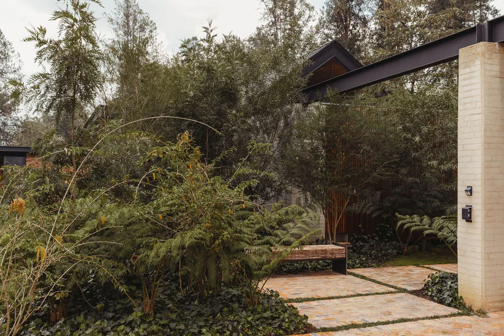 A stone paver path leads past a simple wooden bench and dense green ferns under a dark metal architectural beam and light brick pillar.