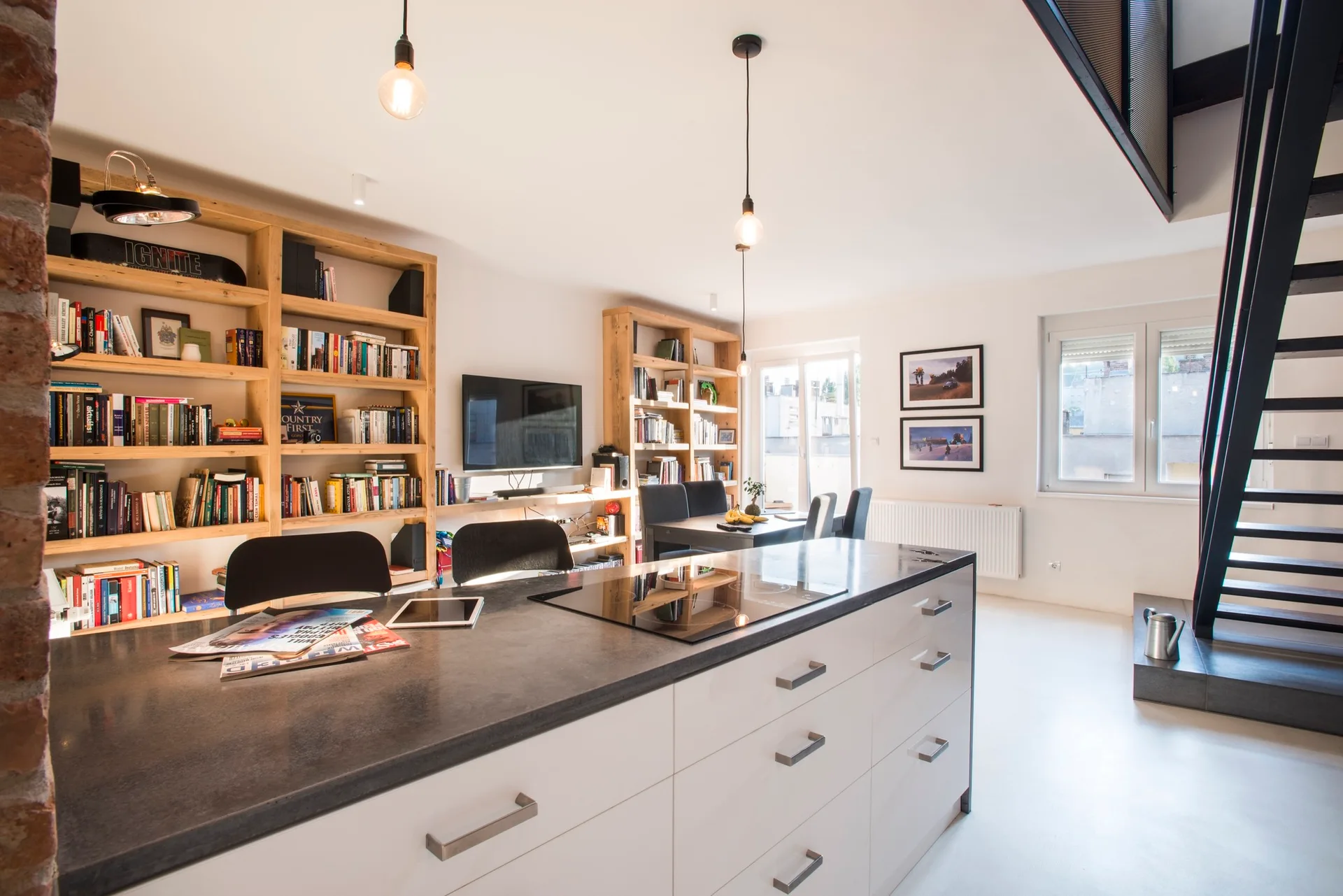 An open-concept kitchen features a dark grey island, floor-to-ceiling wooden bookshelves, white floors, and a black steel staircase.
