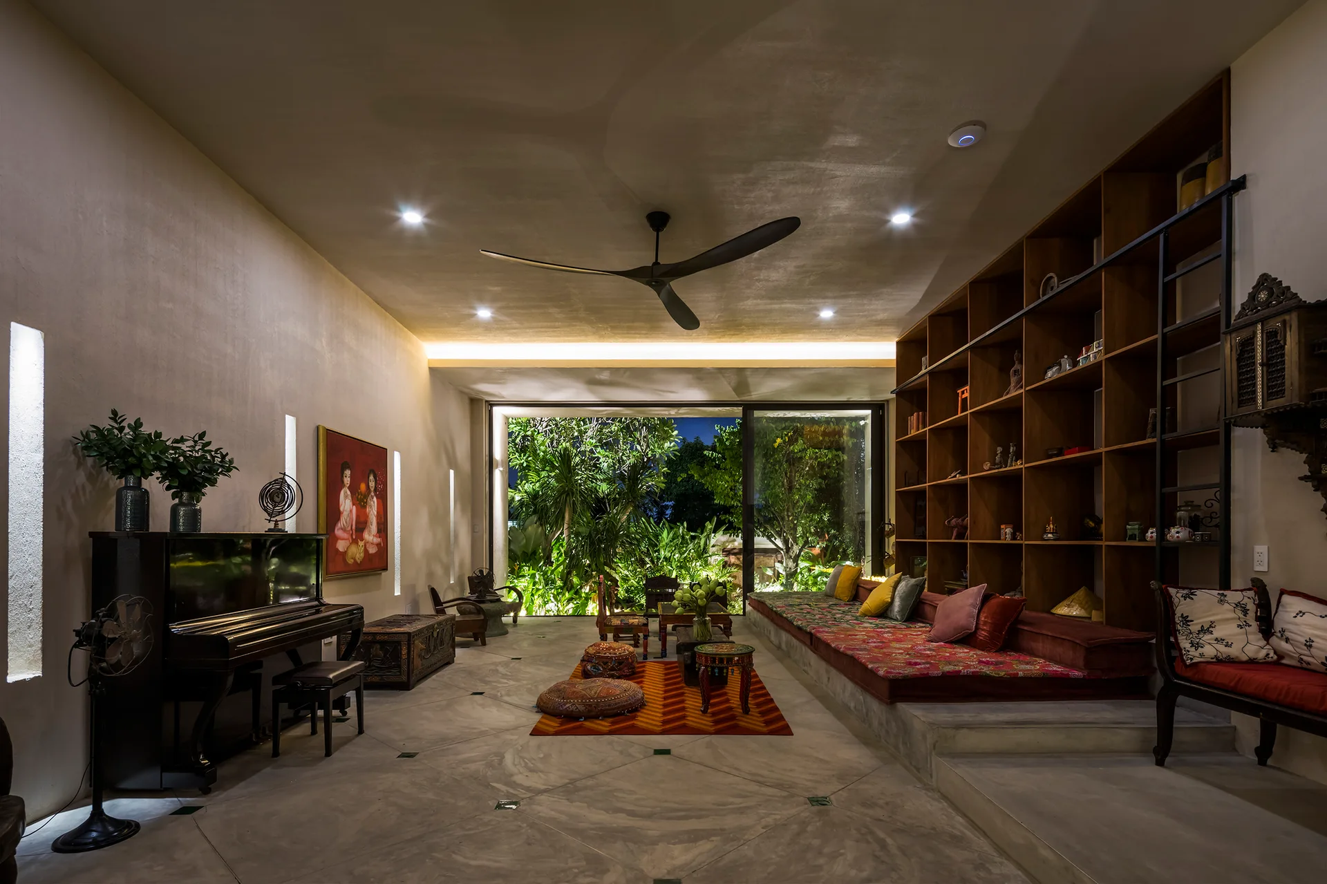 An eclectic living room features a raised platform with colorful cushions, a large built-in wooden shelving unit, and a black upright piano.