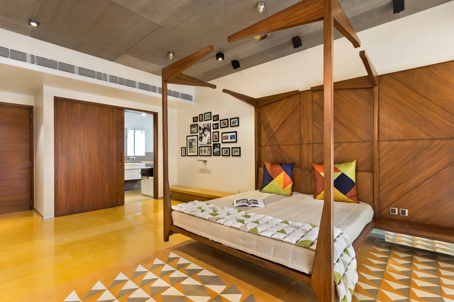 A contemporary bedroom featuring a wooden four-poster bed set against geometric wall paneling and polished yellow flooring with patterned tile accents.