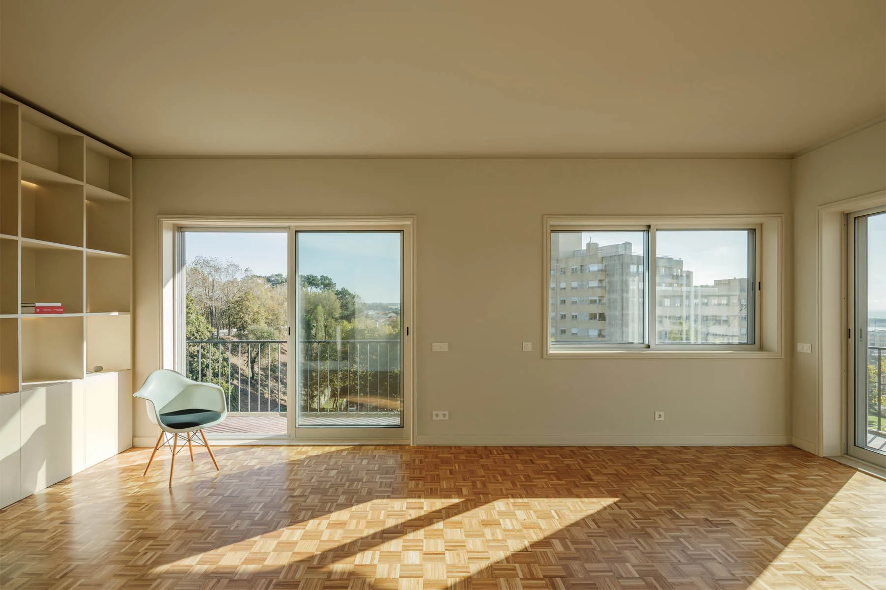 A bright living room featuring basketweave parquet wood flooring, matte beige walls with built-in shelves, and a modern shell chair.