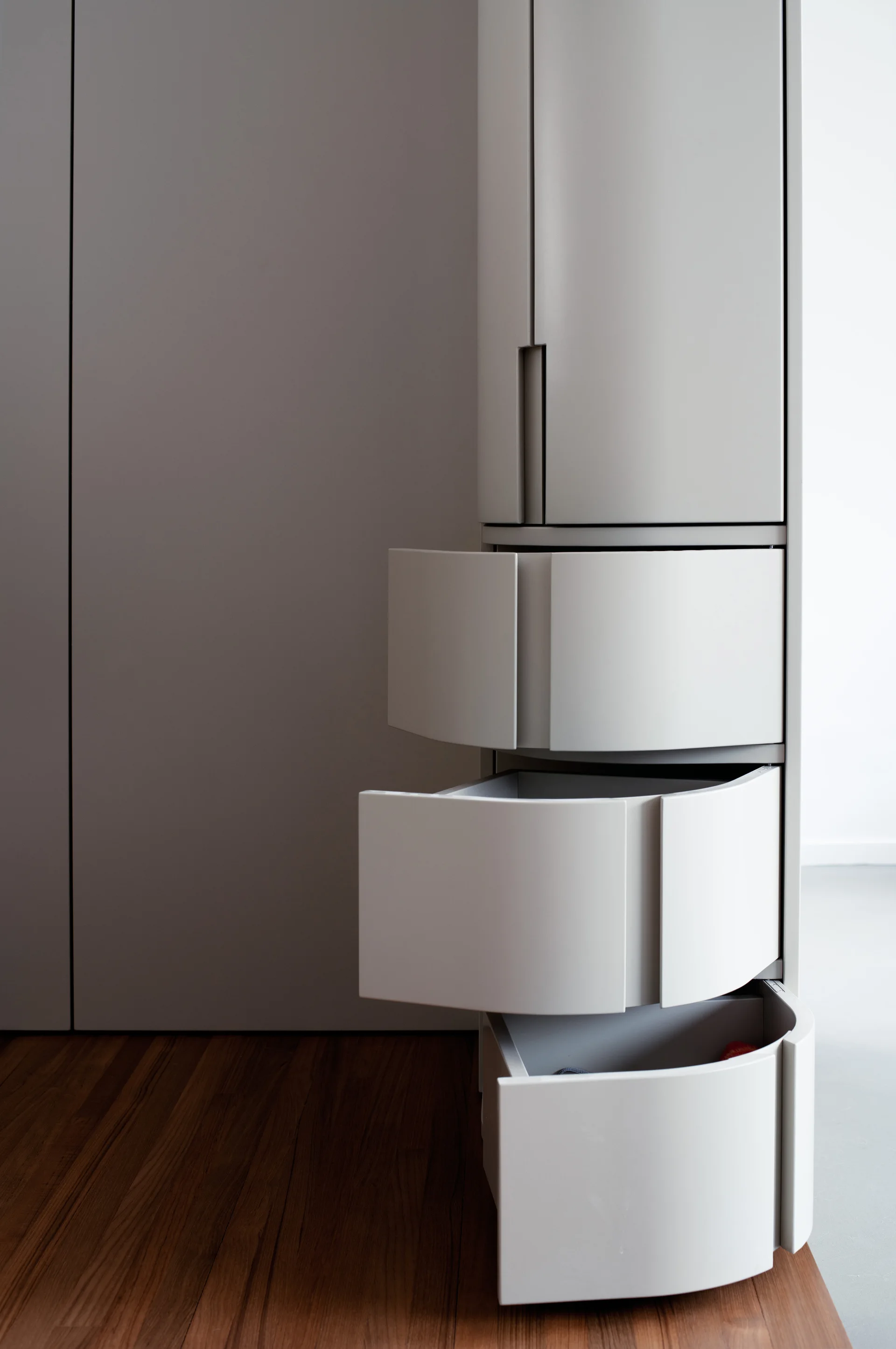 A close-up of modern matte grey cabinetry featuring three open curved drawers set over warm wood plank flooring.