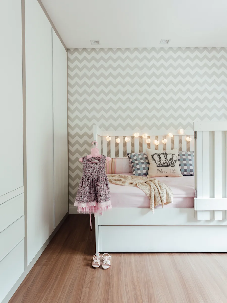 A children's bedroom features a white crib bed set against a beige chevron patterned wall, next to a built-in white wardrobe and wood flooring.