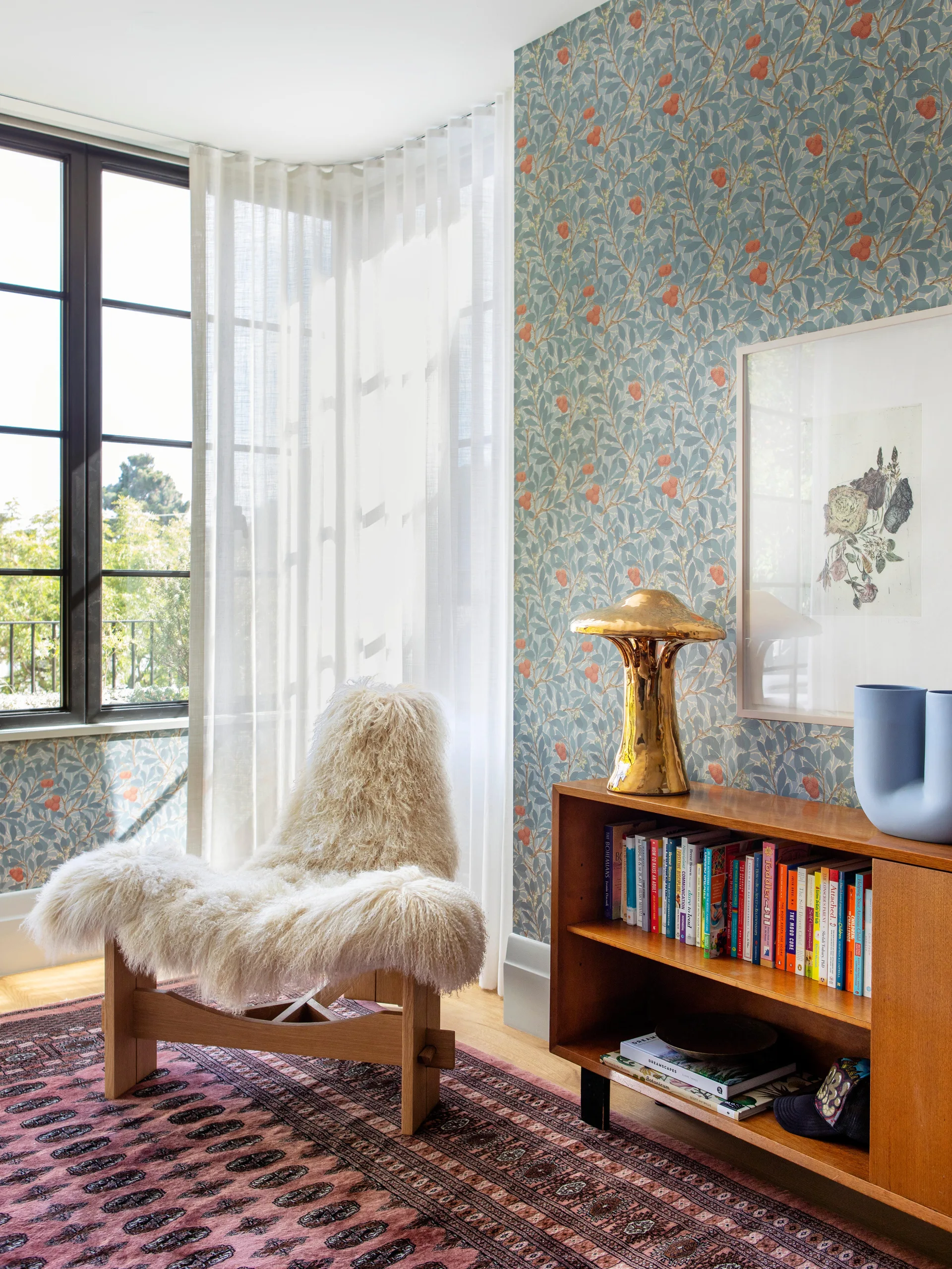 An accent chair draped in fluffy white sheepskin sits next to a low wooden bookshelf in front of blue botanical patterned wallpaper.