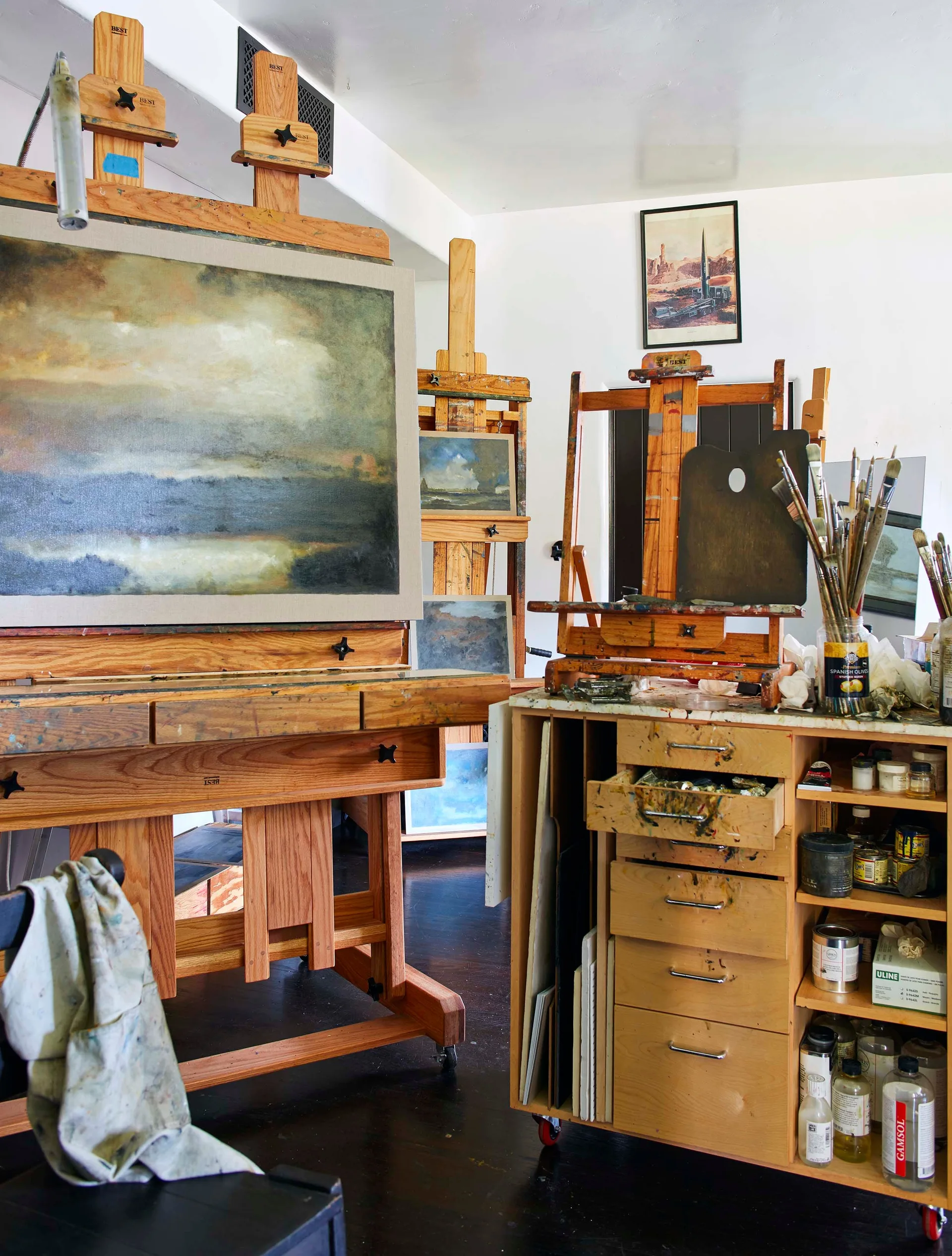 Large wooden easels and a wheeled utility cabinet filled with paintbrushes and art supplies stand on a dark floor in a bright studio.