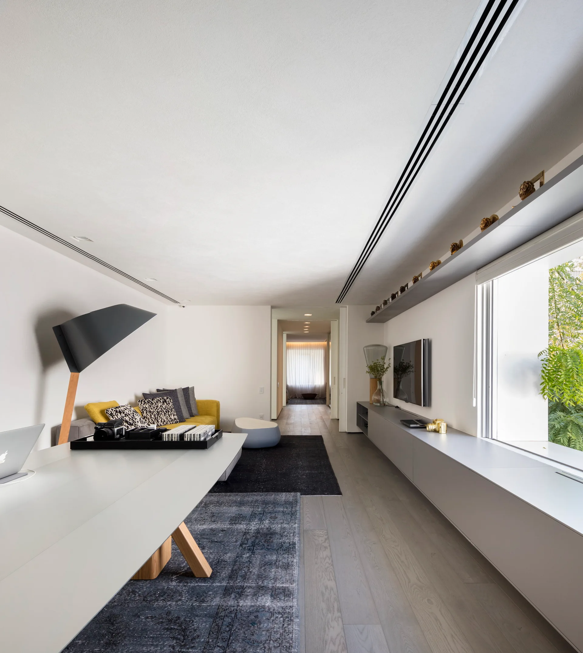 A modern living room features a yellow sofa, a very long grey built-in storage console under a window, and a white desk over grey wood flooring.