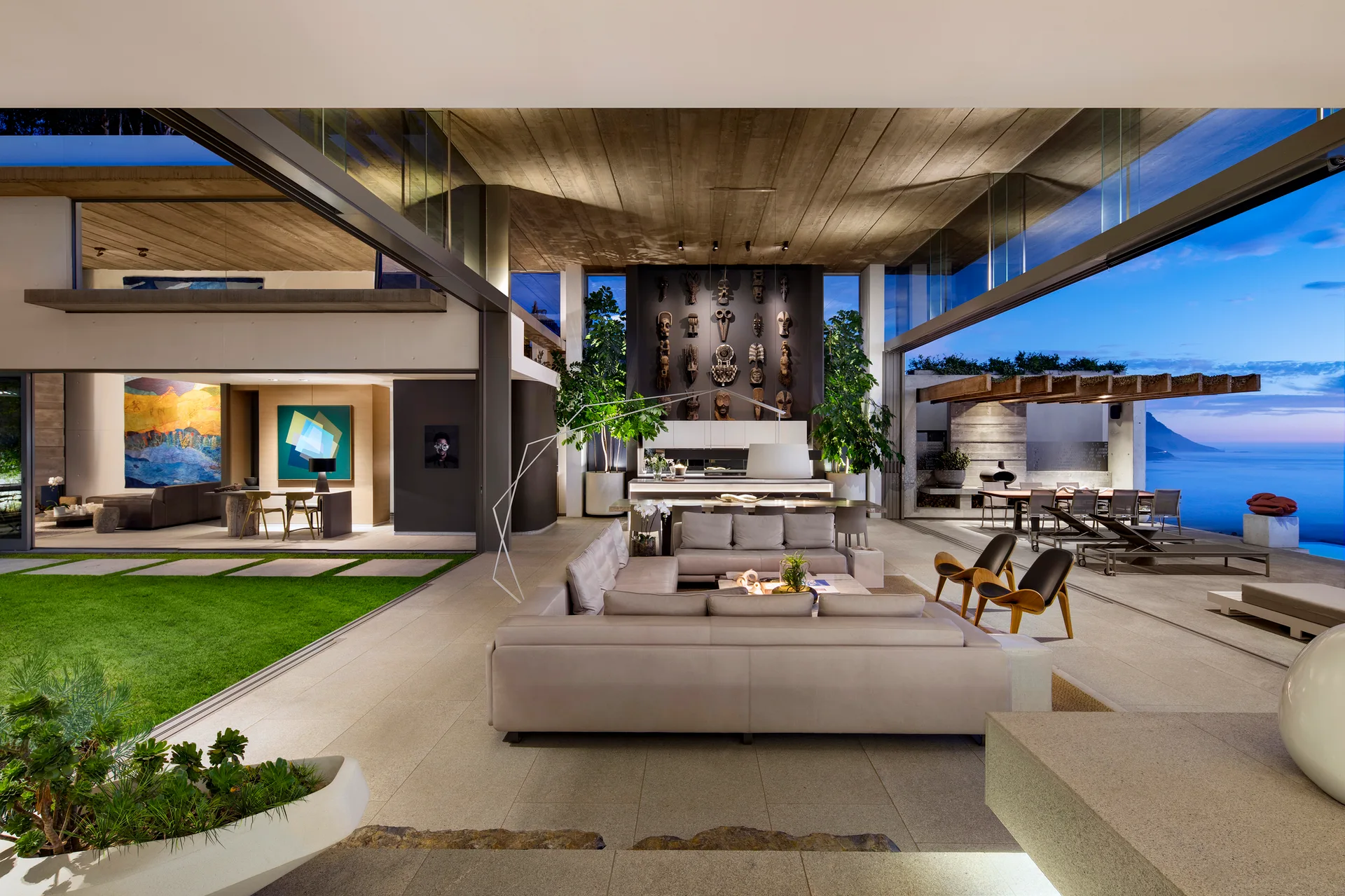 A modern living room with retractable glass walls opening to an outdoor stone patio with ocean views, featuring a modular sofa and wood ceiling.