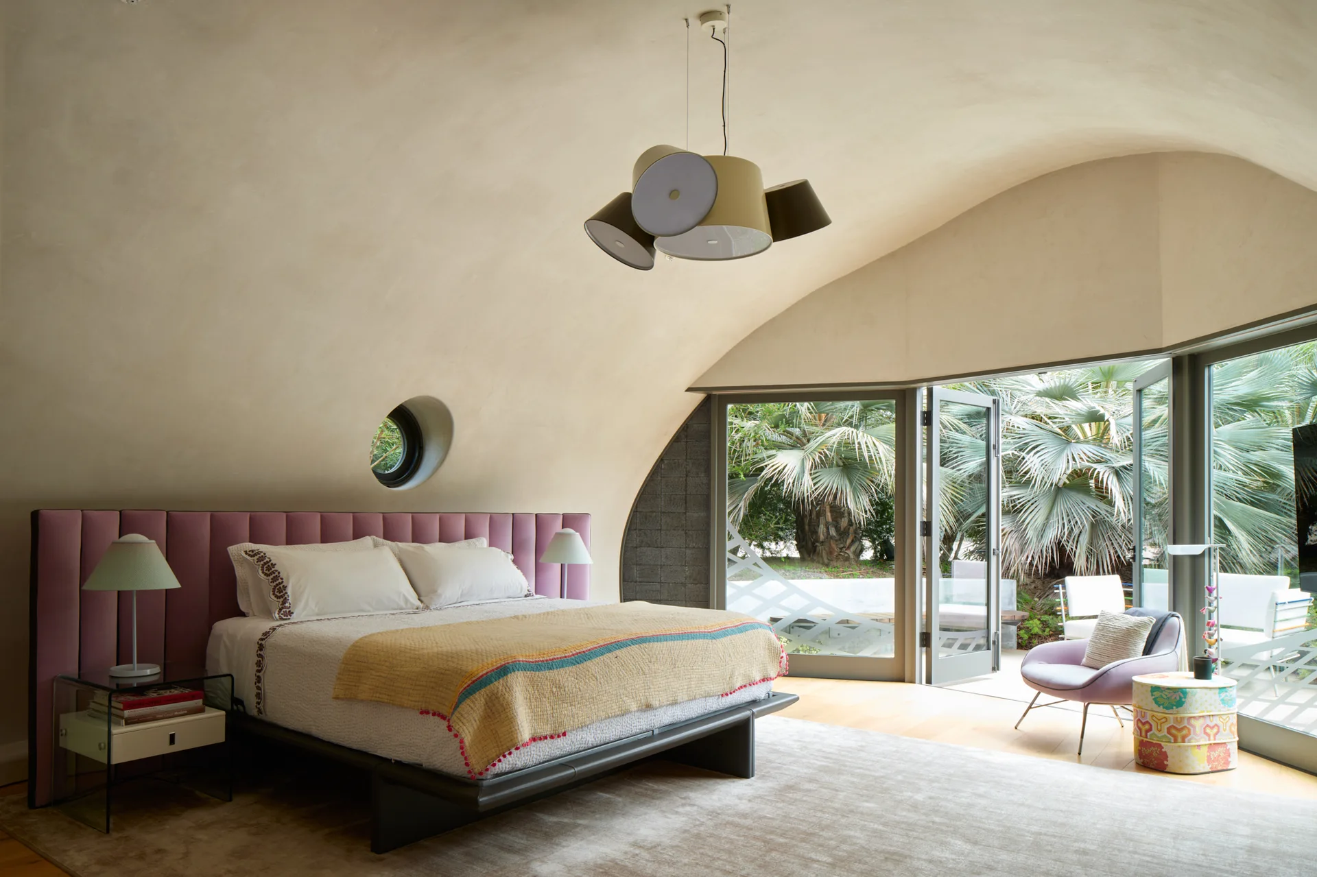 A bedroom with a curved plaster ceiling, a pink upholstered channel headboard, and folding glass doors leading to outdoor tropical plants.