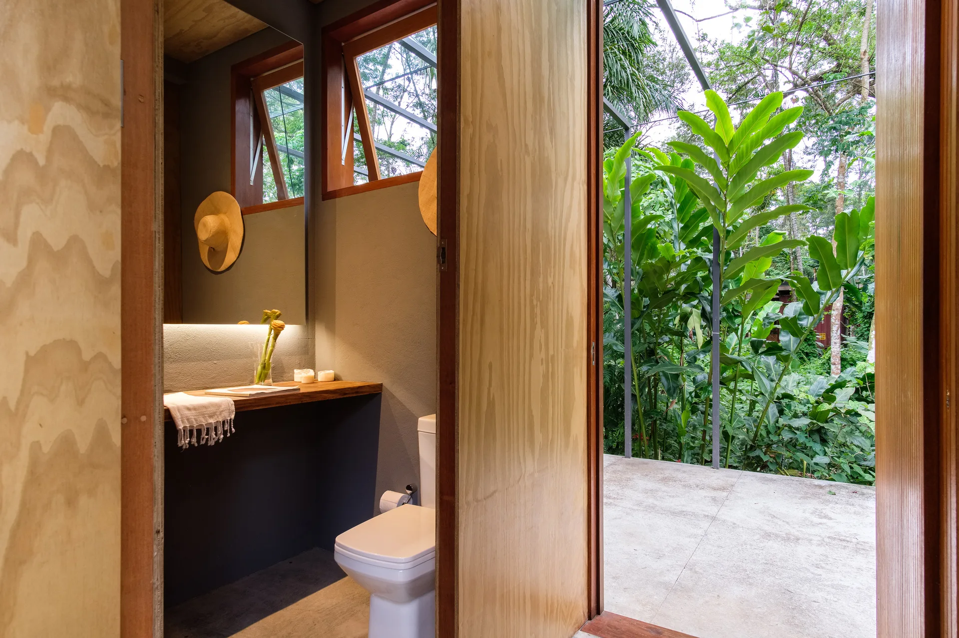 A minimalist bathroom features a floating wooden vanity, a large backlit mirror, and a wide open doorway looking out onto lush green garden foliage.