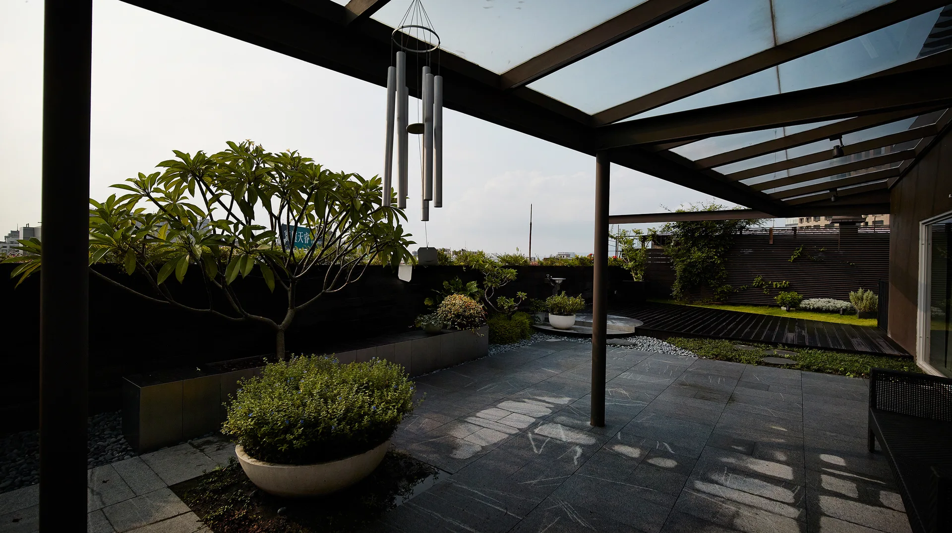 A modern terrace features a metal-framed glass pergola over large grey stone floor tiles, decorated with potted green shrubs and a hanging wind chime.
