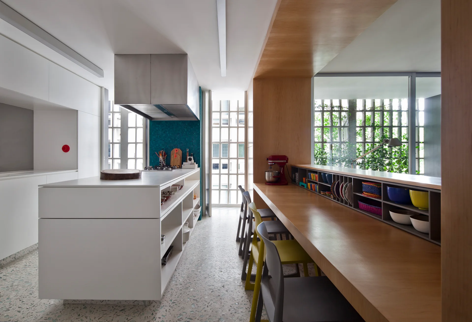 A modern kitchen featuring a white island with open shelving, a long wood breakfast bar with chairs, and a speckled terrazzo floor.