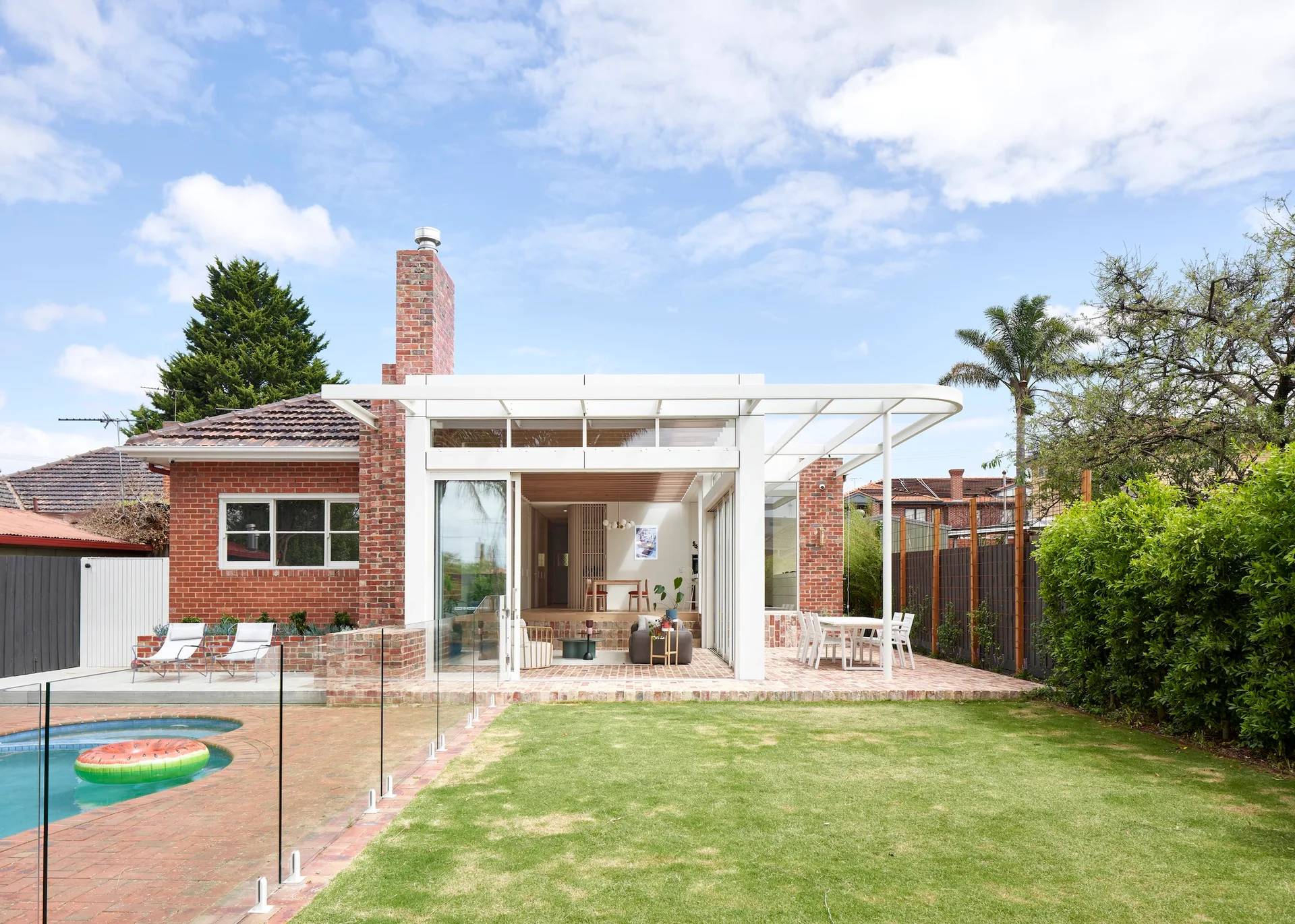 A red brick house features a modern white pergola extension opening onto a paved brick patio, green lawn, and swimming pool.