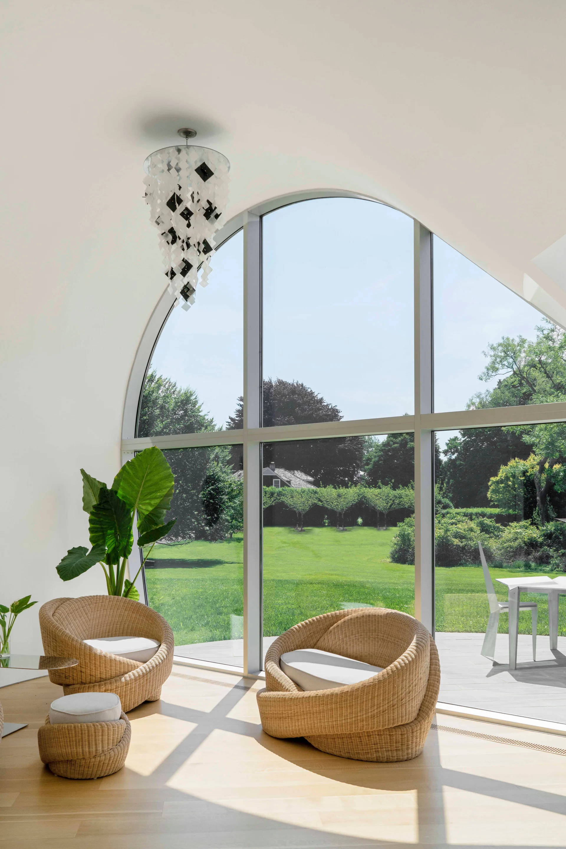 Two rounded rattan armchairs face a large arched floor-to-ceiling window overlooking a green lawn in a bright room with light wood floors.