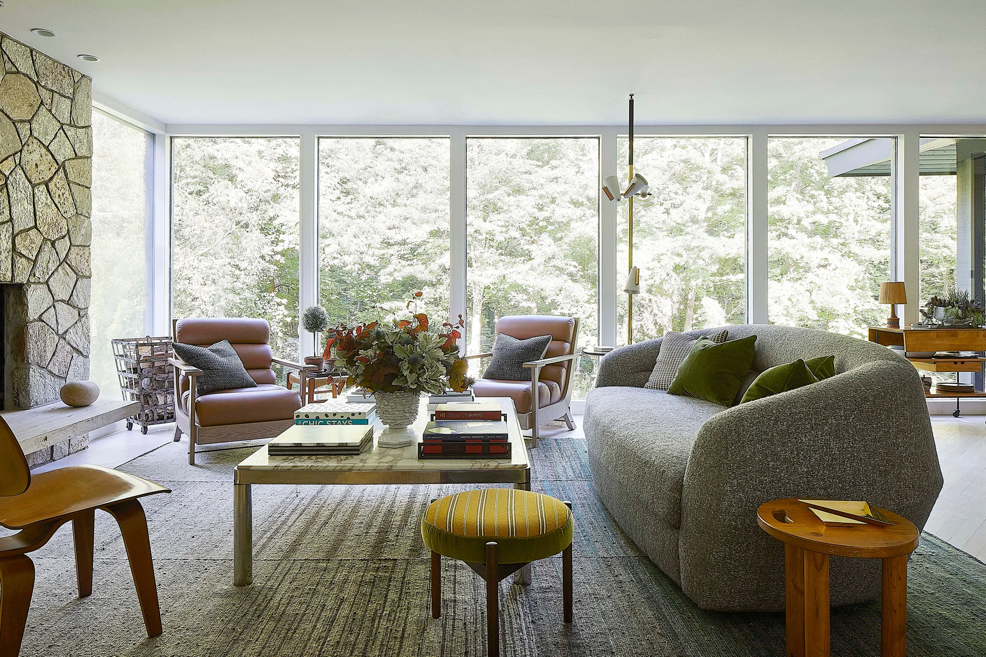 A sunlit living room with a curved grey sofa, a marble coffee table, tan leather armchairs, and a large stone fireplace next to floor-to-ceiling windows.