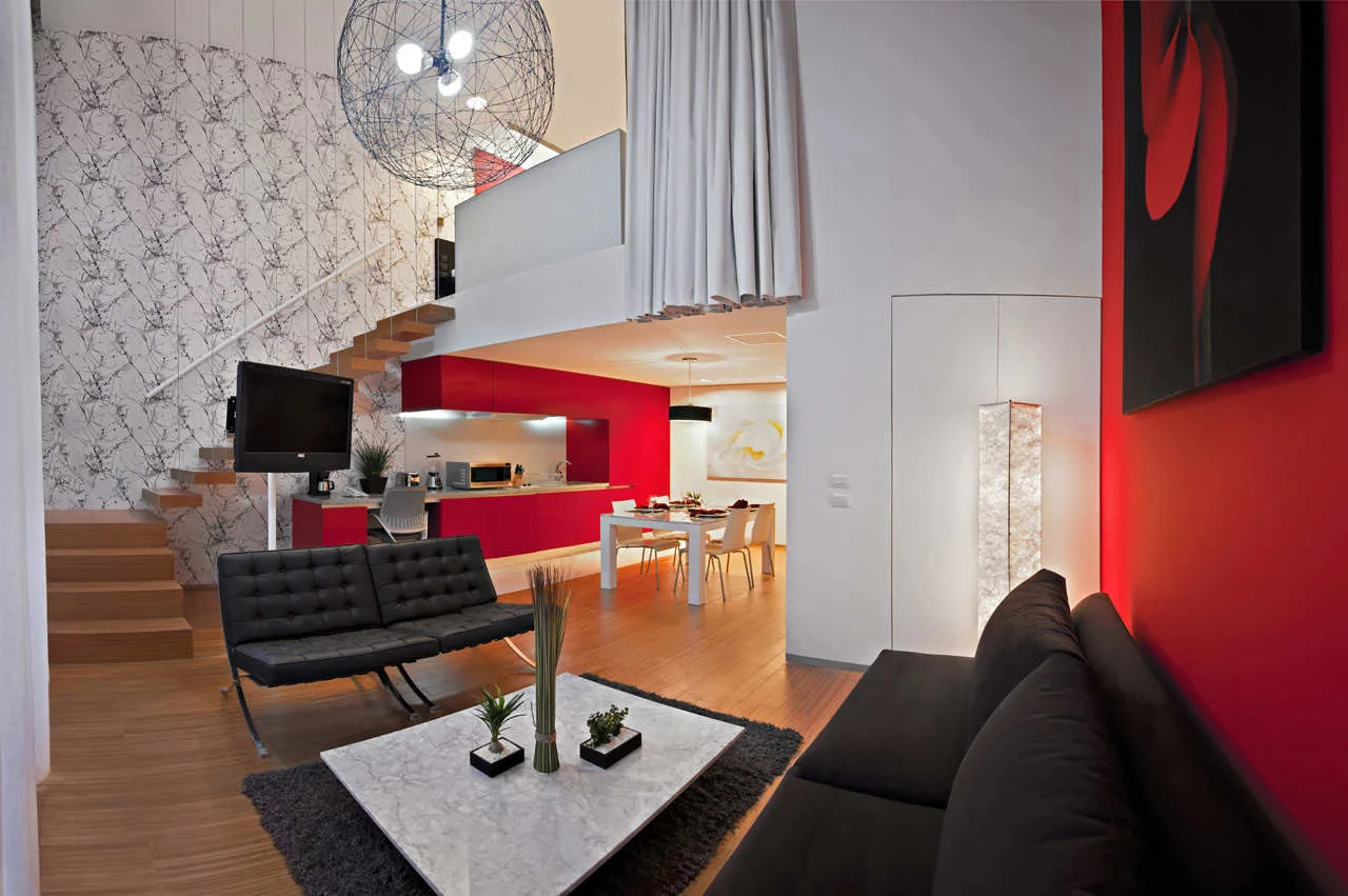 A modern split-level living room features black sofas, a marble coffee table, a floating wooden staircase, and vibrant red accent walls and kitchen cabinets.