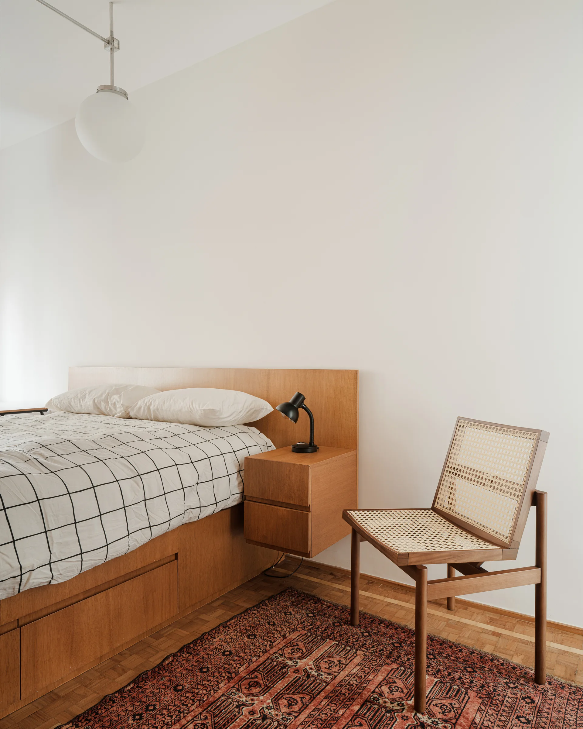 A minimalist bedroom features a wooden platform bed, integrated floating nightstand, grid bedding, and a cane chair on a patterned rug.