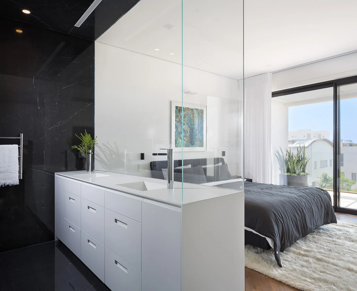 A modern bedroom features a bed with dark linens, a white shag rug, and an ensuite vanity area separated by a tall glass partition.