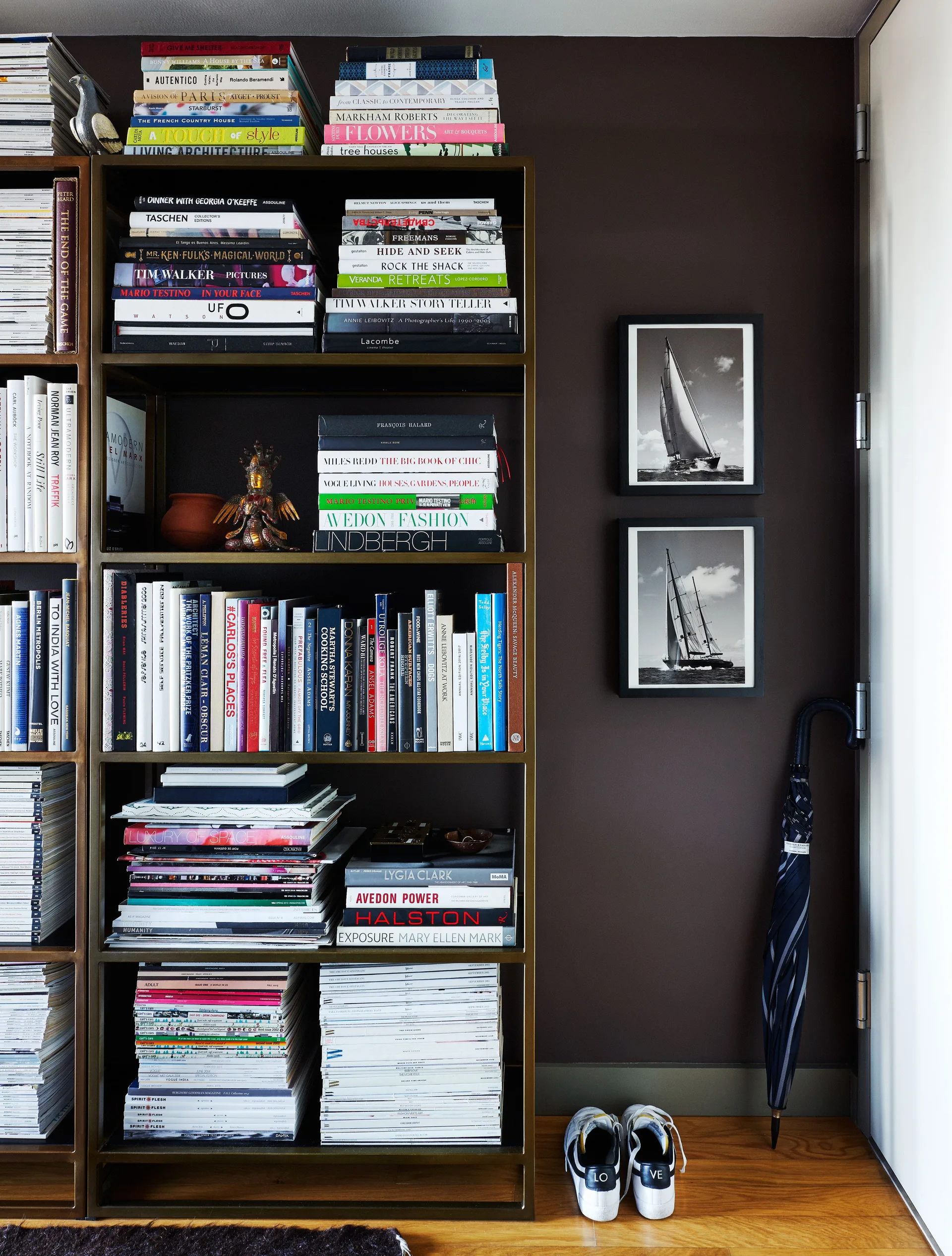 A brass bookshelf filled with books stands against a dark brown wall next to stacked framed sailboat prints and white sneakers on wood flooring.