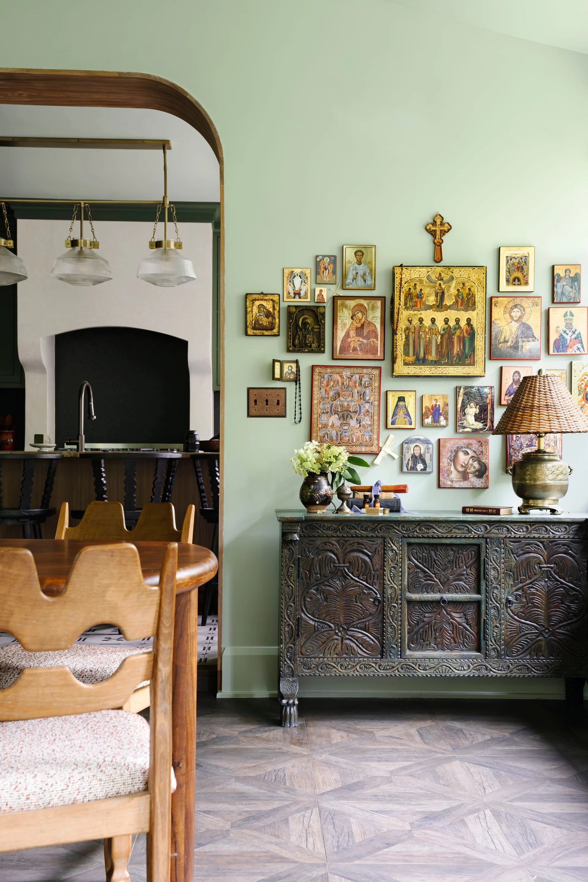 A dining room features a carved wooden credenza against a sage green wall decorated with a gallery of religious icons next to a wood-trimmed archway.