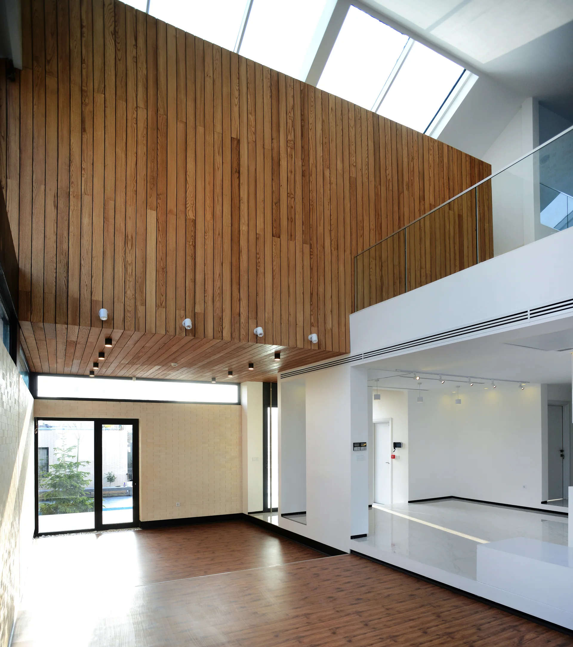 A modern multi-level interior featuring a tall vertical wood-paneled wall, overhead skylights, white walls, and a glass-railed mezzanine.