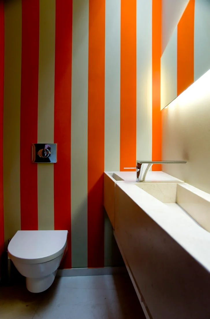 A modern powder room features bold orange and beige striped wallpaper, a wall-mounted toilet, and a long integrated stone sink vanity.