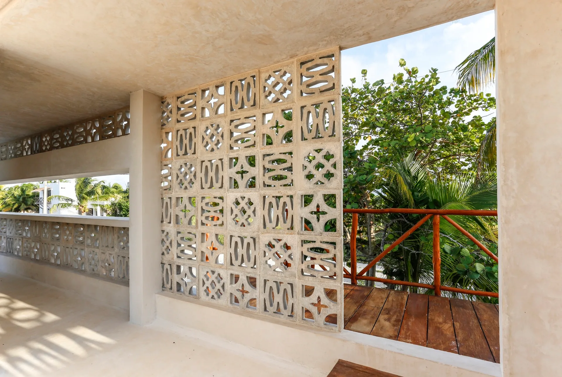 A covered concrete terrace features a decorative geometric breeze block screen wall next to a wooden deck with a timber railing overlooking lush trees.