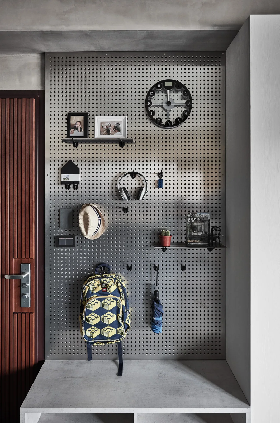 A metallic silver pegboard wall panel hangs above a concrete bench in an entryway, holding a backpack, clock, headphones, and small shelves.
