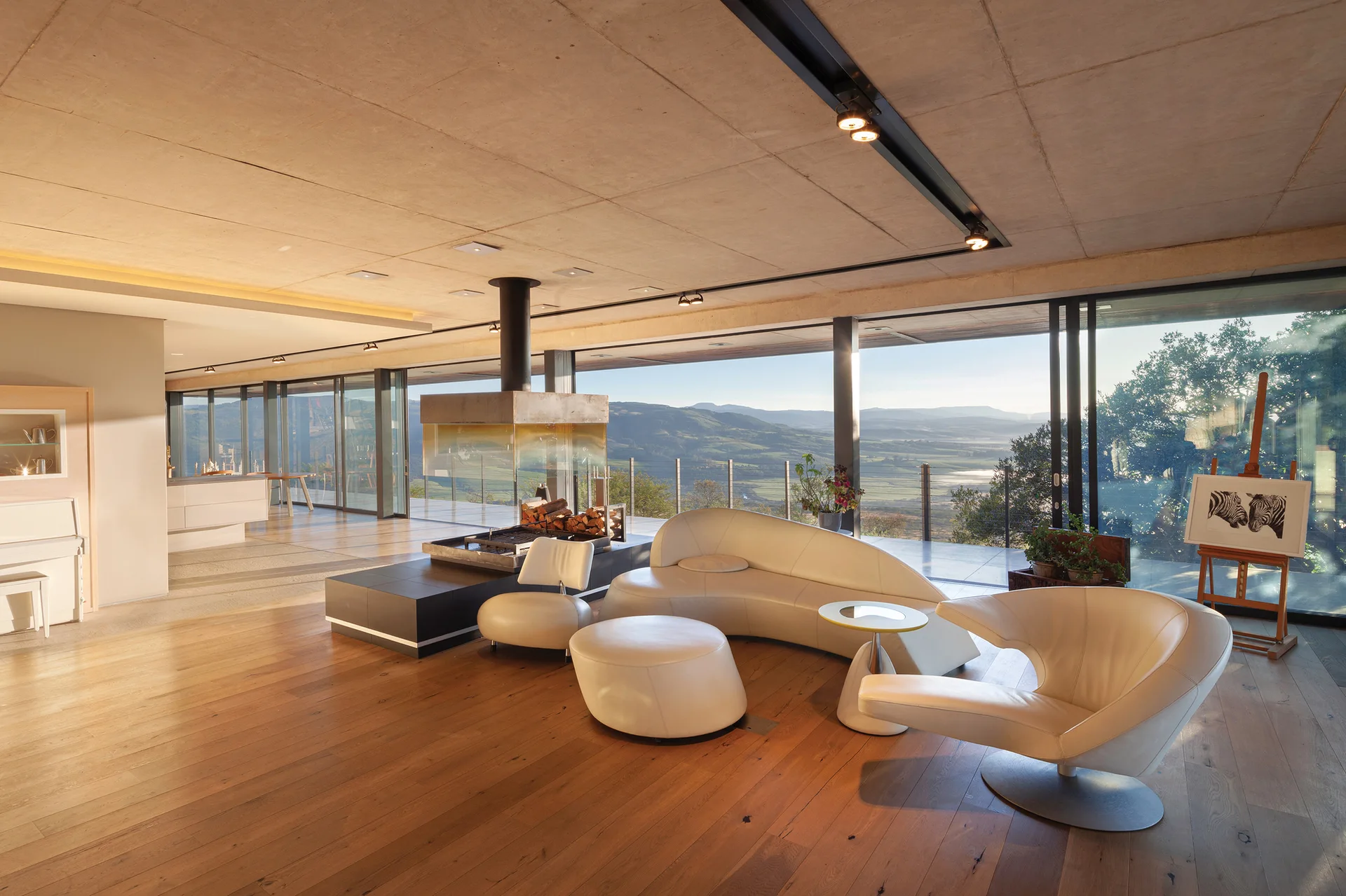 A modern living room features a curved cream leather sofa and a central glass fireplace set against a wall of floor-to-ceiling windows.