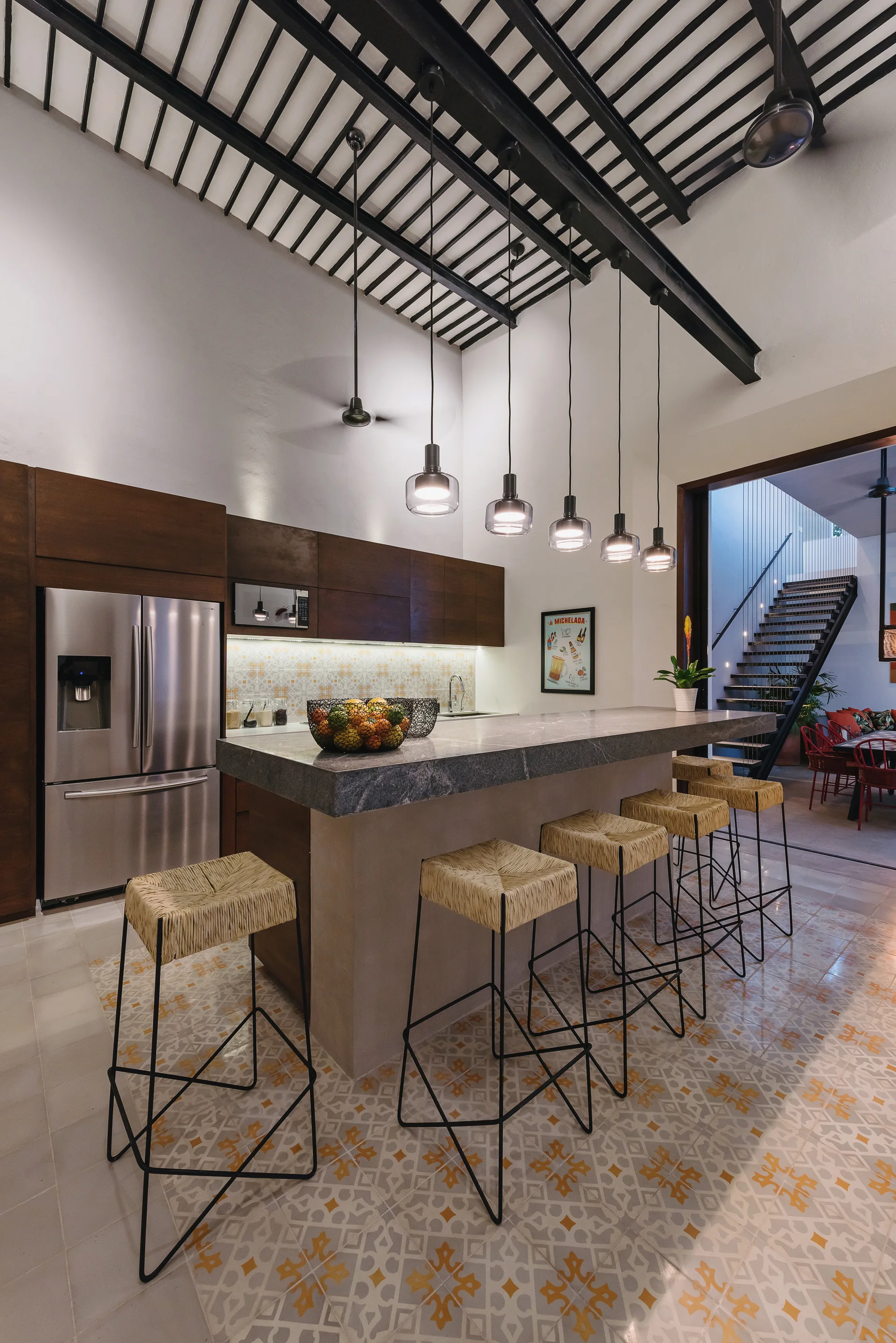 A modern kitchen featuring a stone-topped island with woven bar stools, five glass pendant lights, and exposed black ceiling beams.