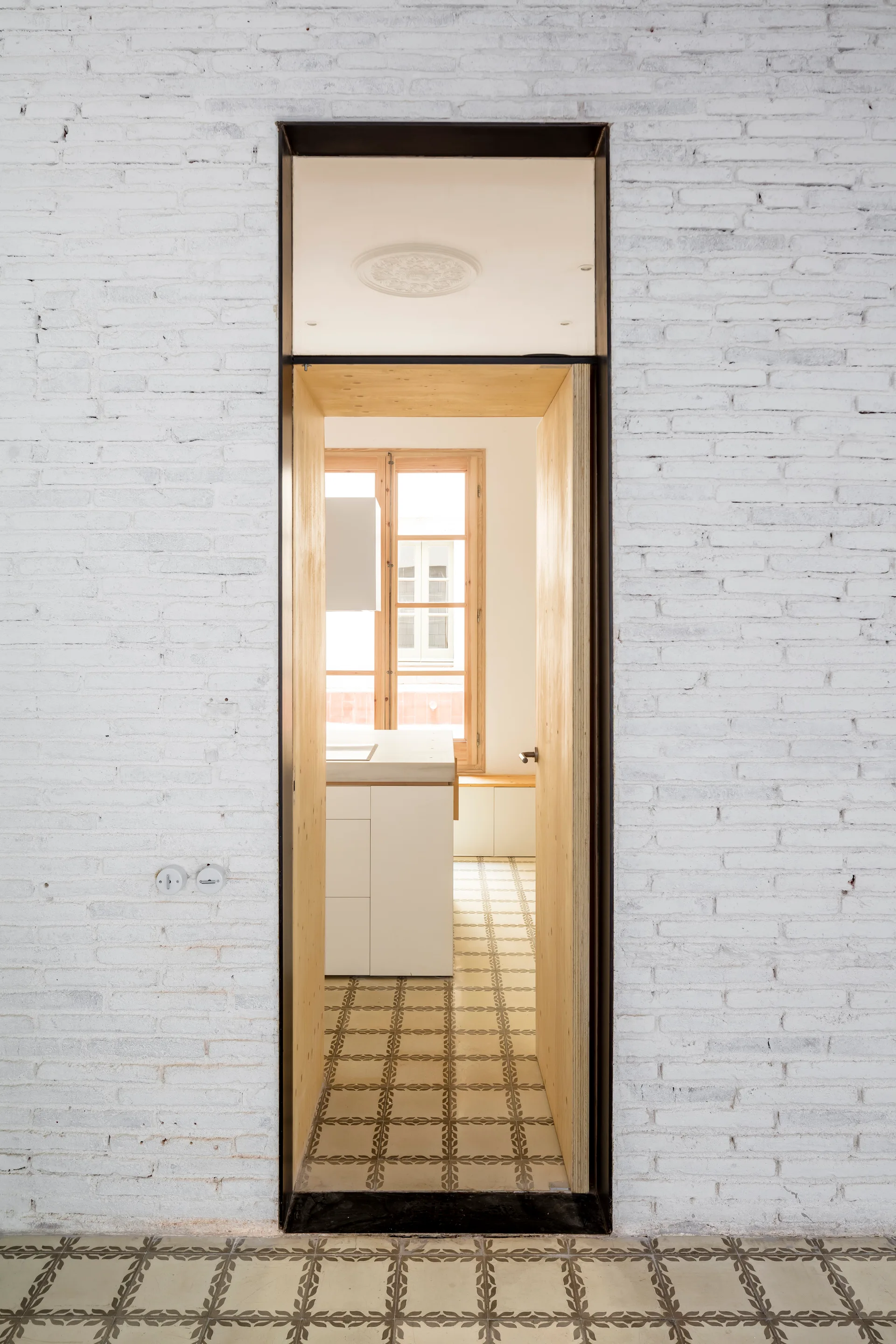 A view through a deep natural wood cased doorway set in a white painted brick wall with patterned floor tiles leading into a sunlit kitchen.