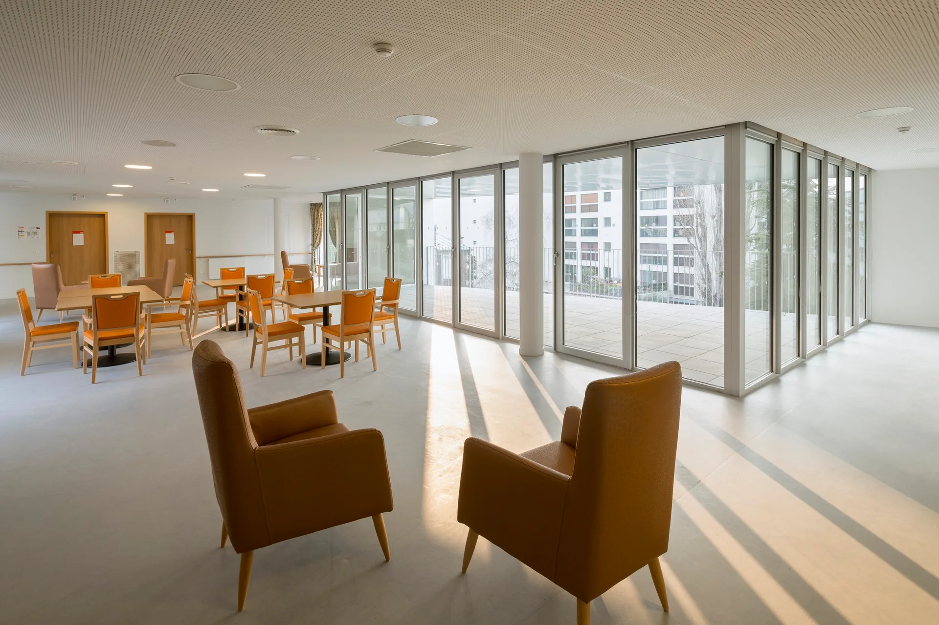 Two brown armchairs stand in the foreground of a sunlit common room featuring several wooden tables with orange chairs and large glass walls.
