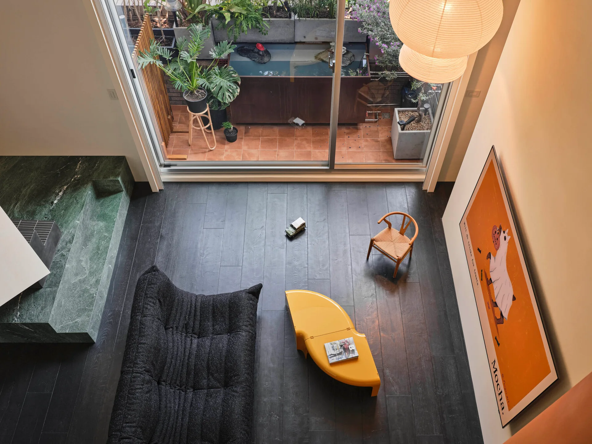 An overhead view of a modern living room featuring a black tufted sofa, a curved yellow coffee table, a green marble hearth, and sliding patio doors.