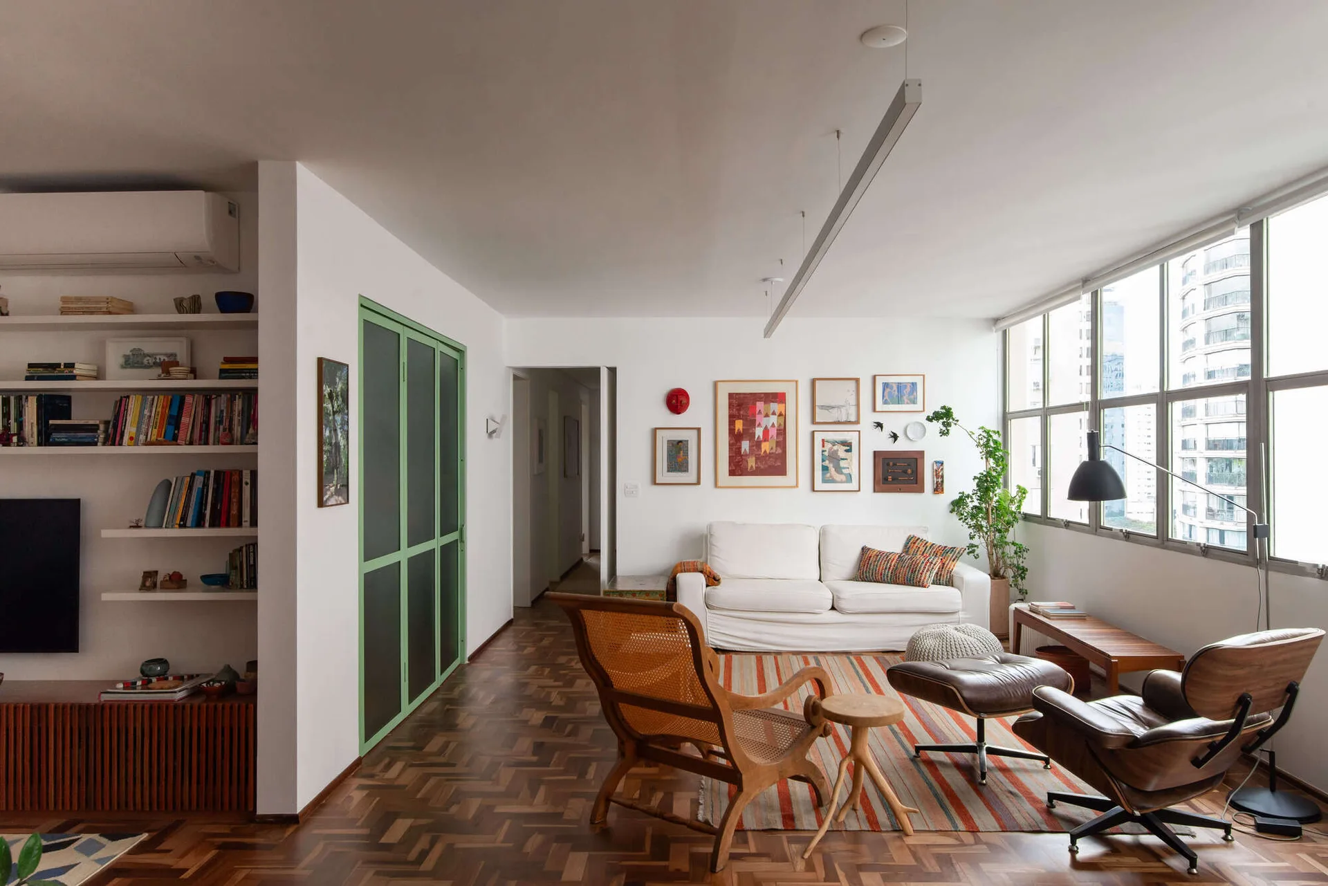 A sunlit living room features parquet wood floors, mid-century modern lounge chairs, a white sofa, and a green-framed sliding door.