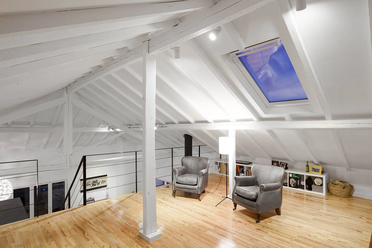 A modern attic loft featuring two grey leather armchairs, light wood floors, and white sloped ceilings with painted exposed beams.