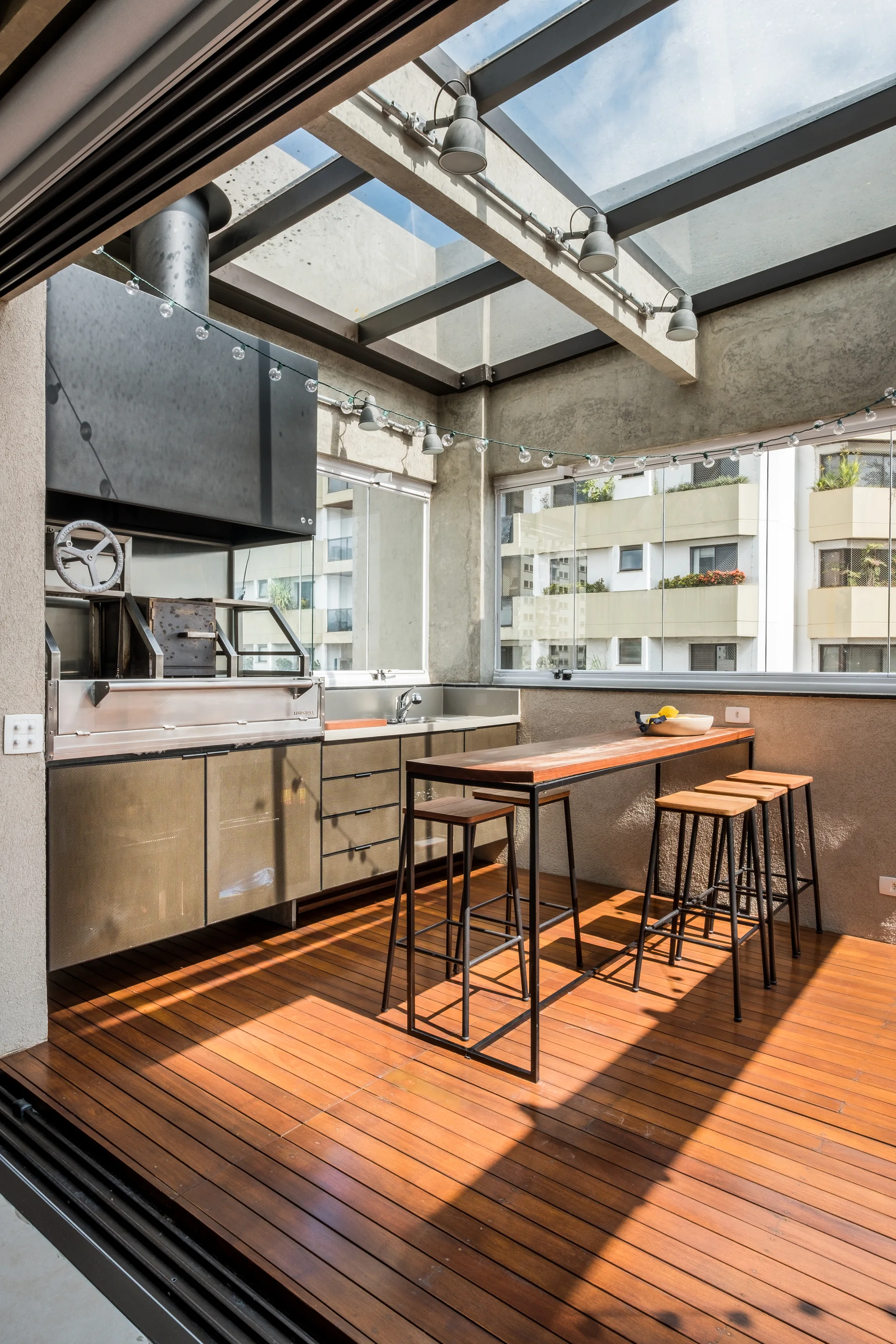 A covered terrace kitchen features a stainless steel grill station, wooden decking, and a glass-paneled roof with concrete beams.